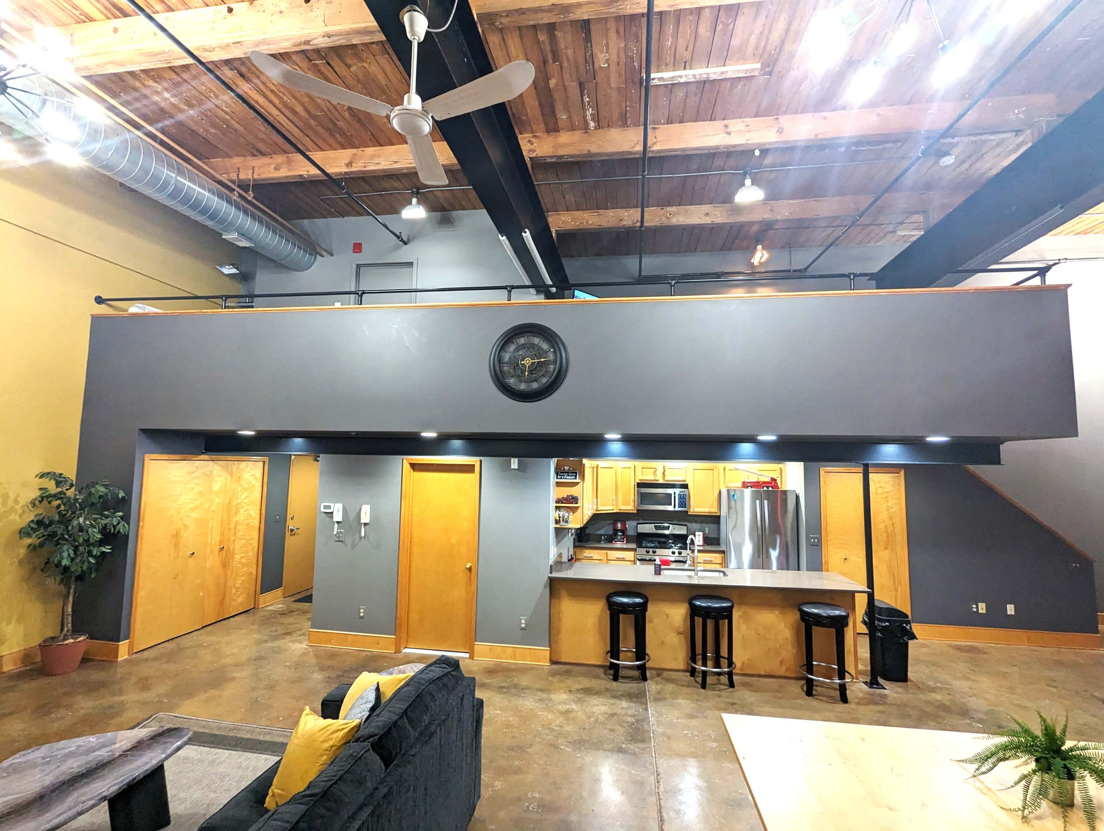 An open-concept loft space with a kitchen, bar stools, and a living area beneath a wooden ceiling.