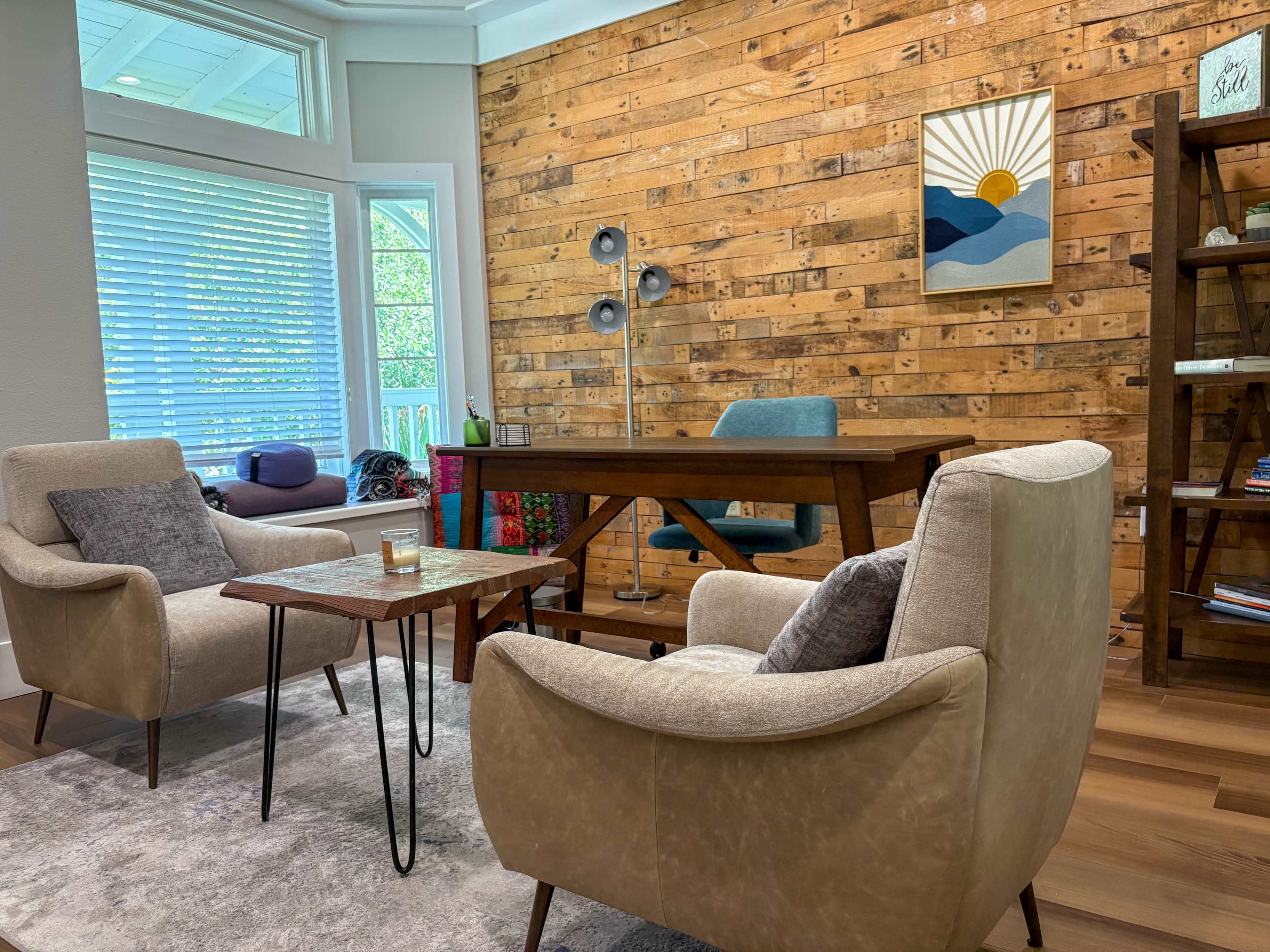 A cozy office space features two armchairs, a wooden table, and a desk against a rustic wood-paneled wall.