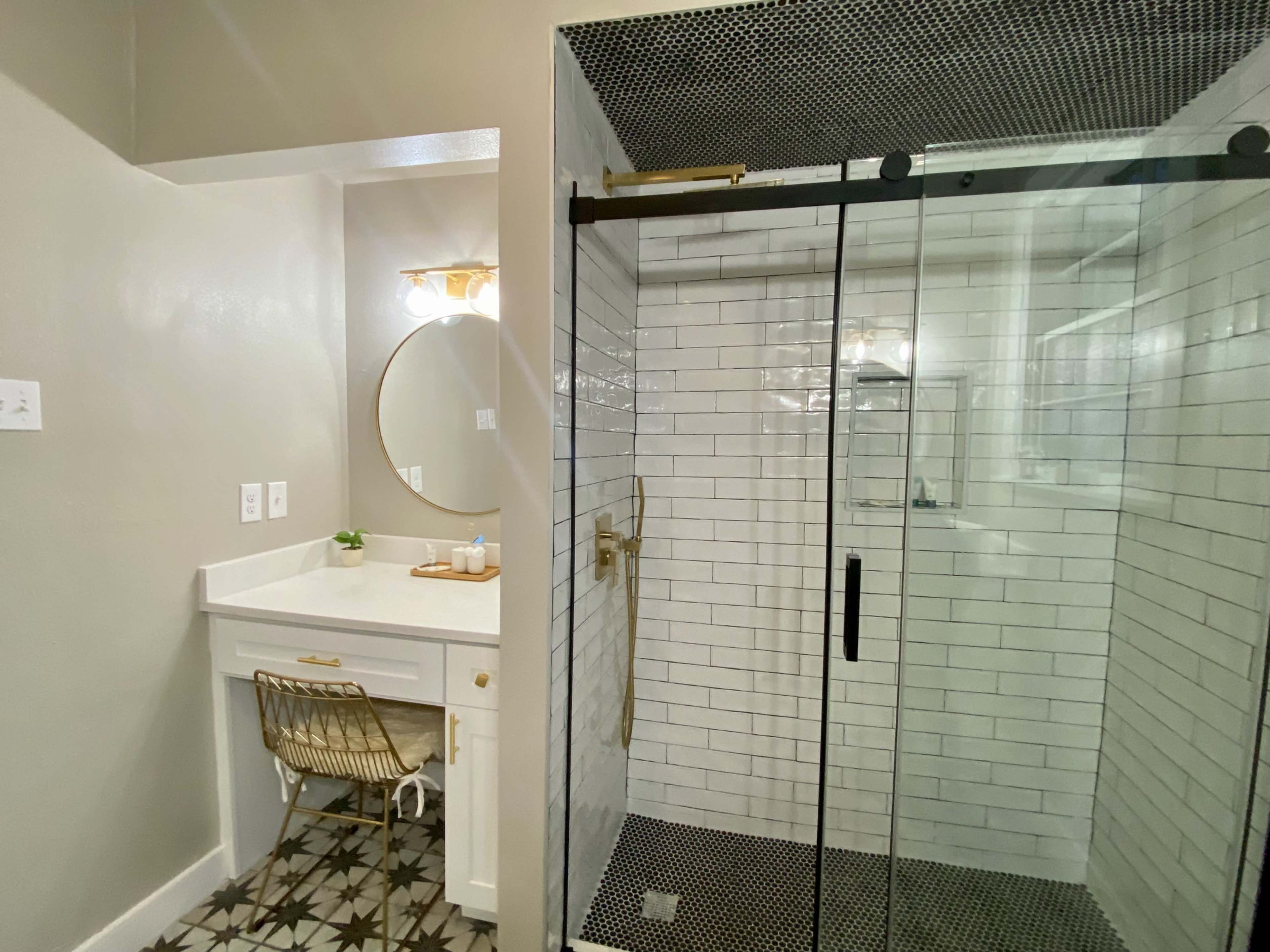 The image shows a bathroom with a glass shower, tiled walls, a round mirror above a sink, and a small desk with a chair.
