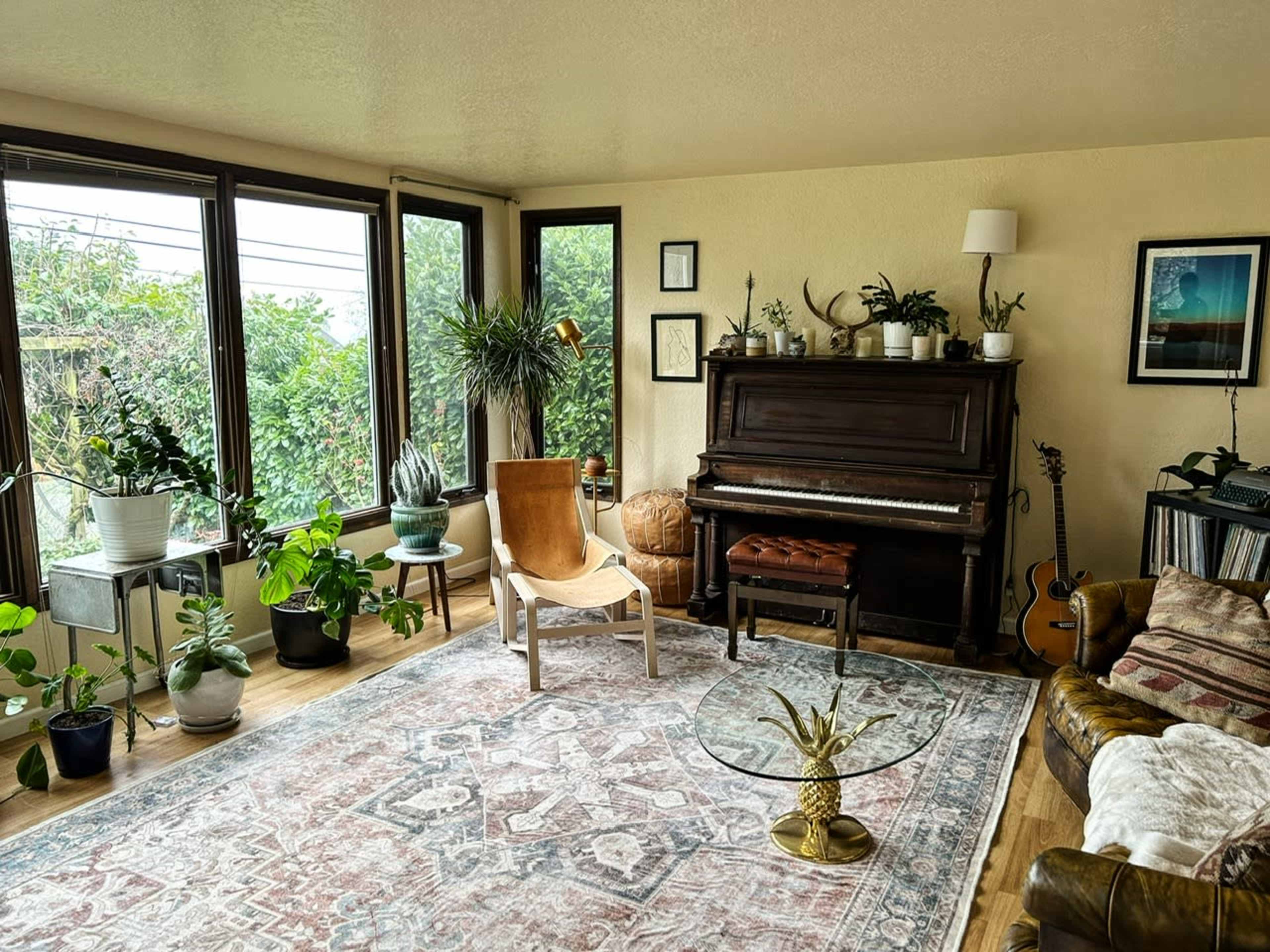 A cozy living room features a piano, several potted plants, a guitar, and a large area rug, with ample natural light coming through the windows.