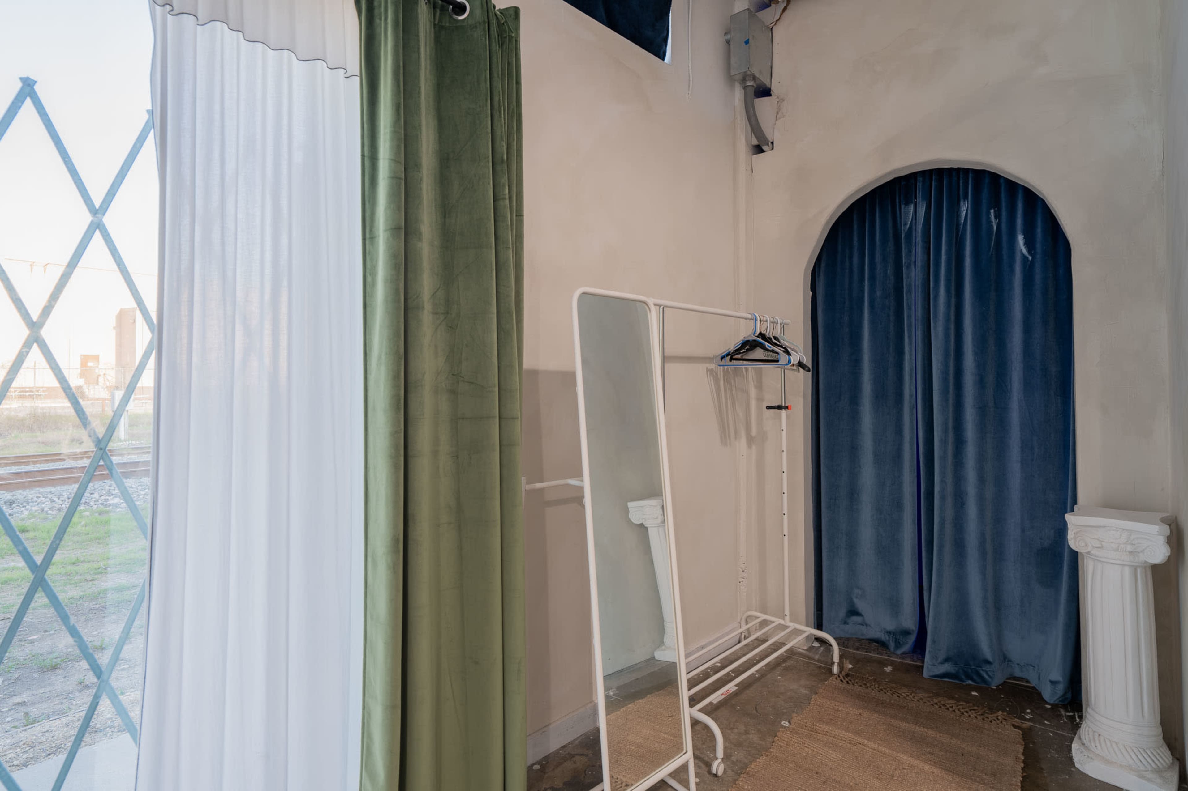 The image shows a corner of a room featuring a curved archway with blue curtains, a clothing rack, and draped fabric in green and white.