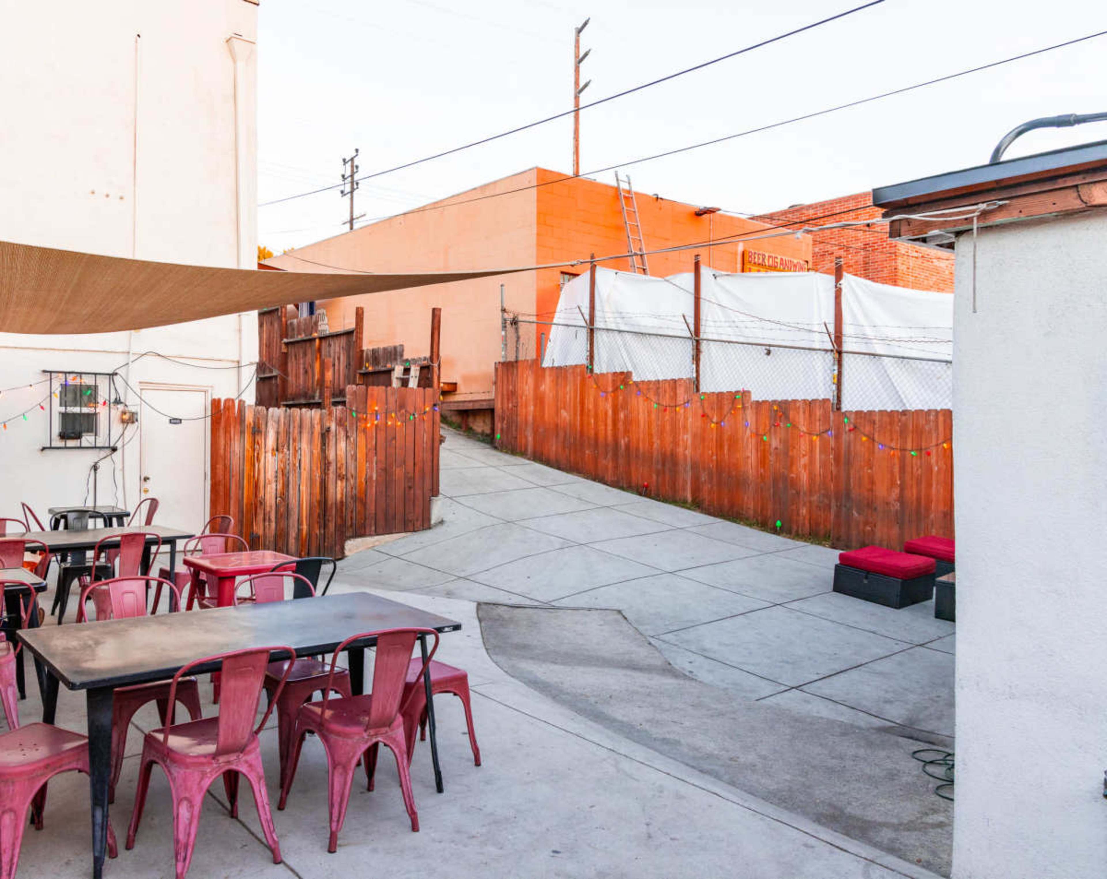 The image shows an outdoor area with several red metal tables and chairs, leading to a pathway bordered by wooden fencing and a white tarp.