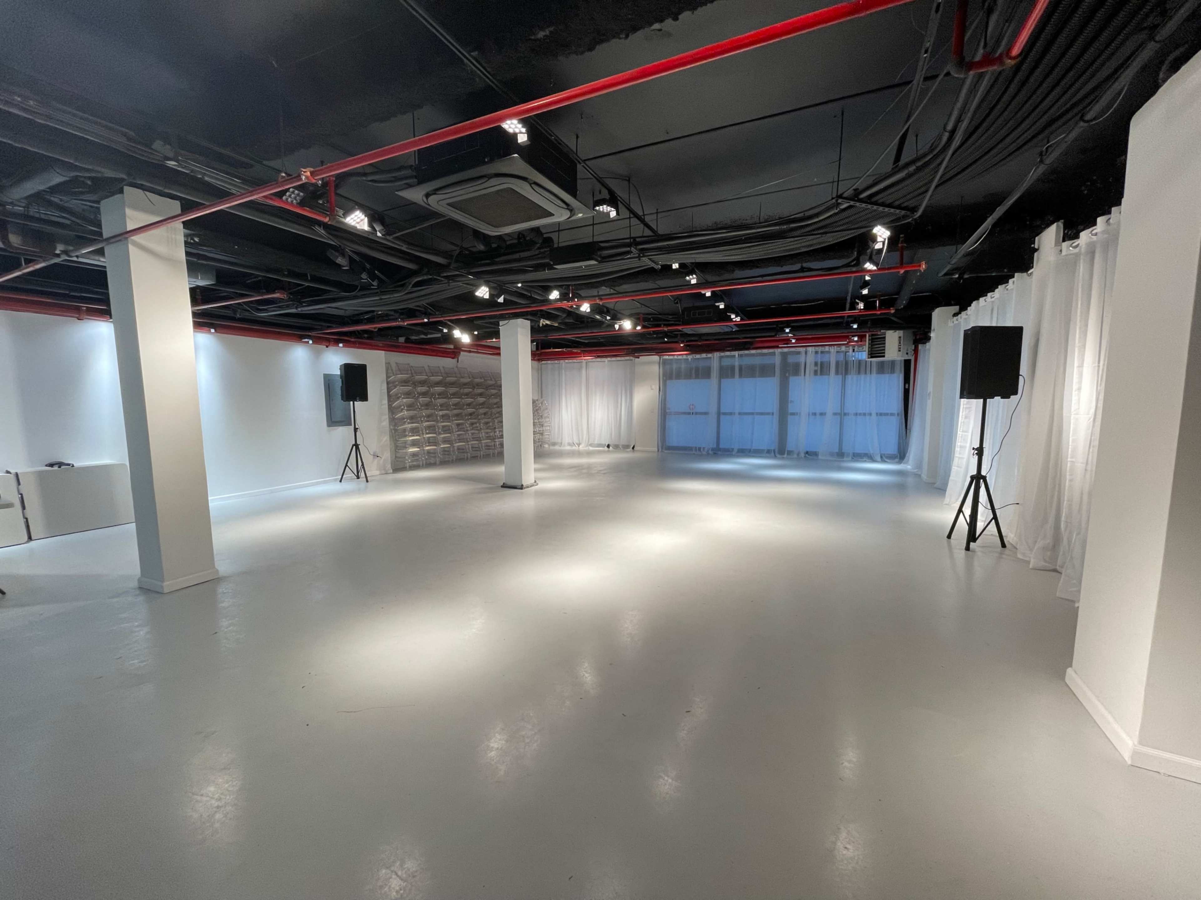 An empty, well-lit room with polished white floors, exposed black ceilings, and large windows letting in natural light.