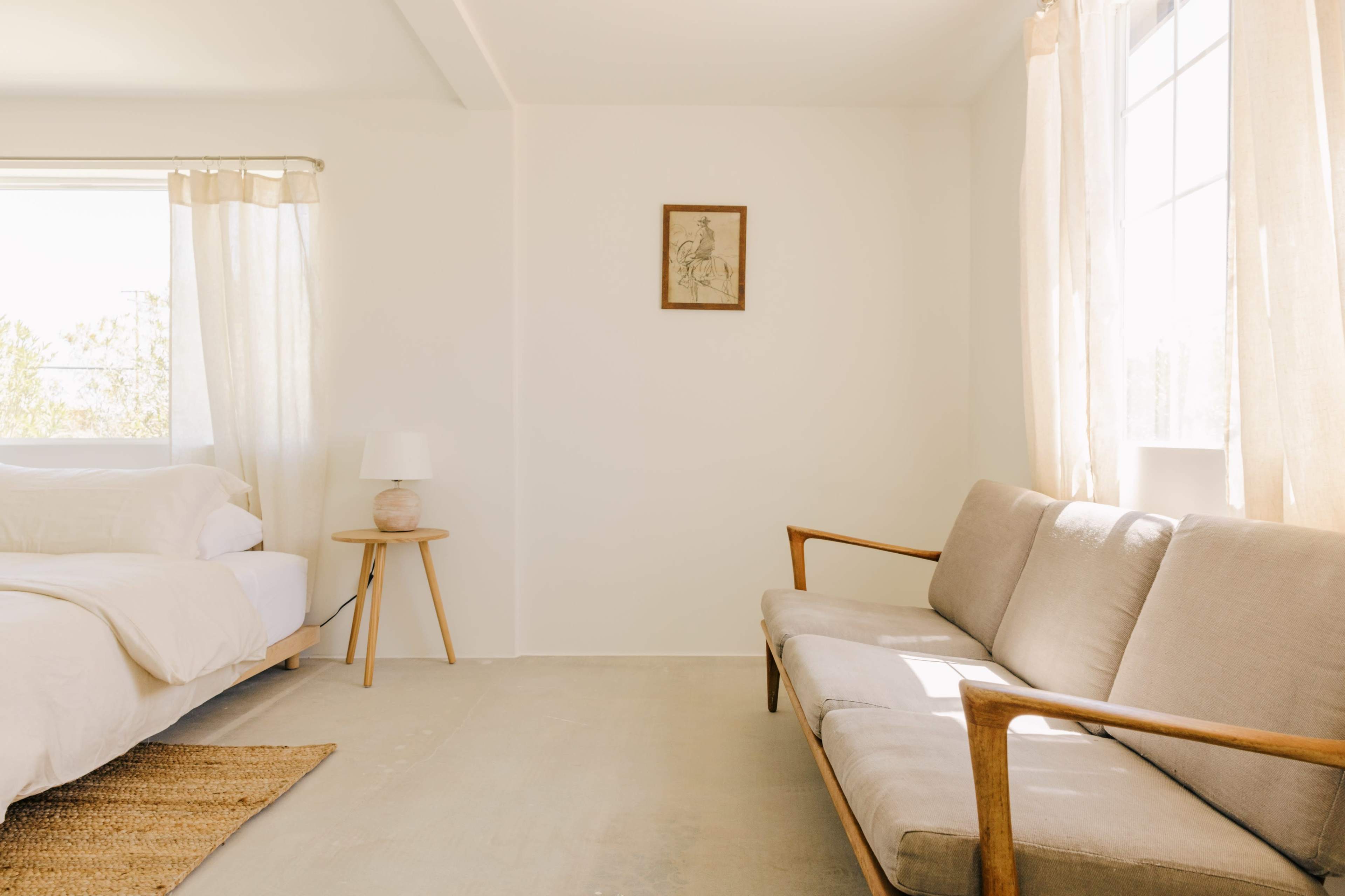 The image shows a minimalistic bedroom with a bed, a light-colored sofa, a lamp, and a small framed picture on the wall.