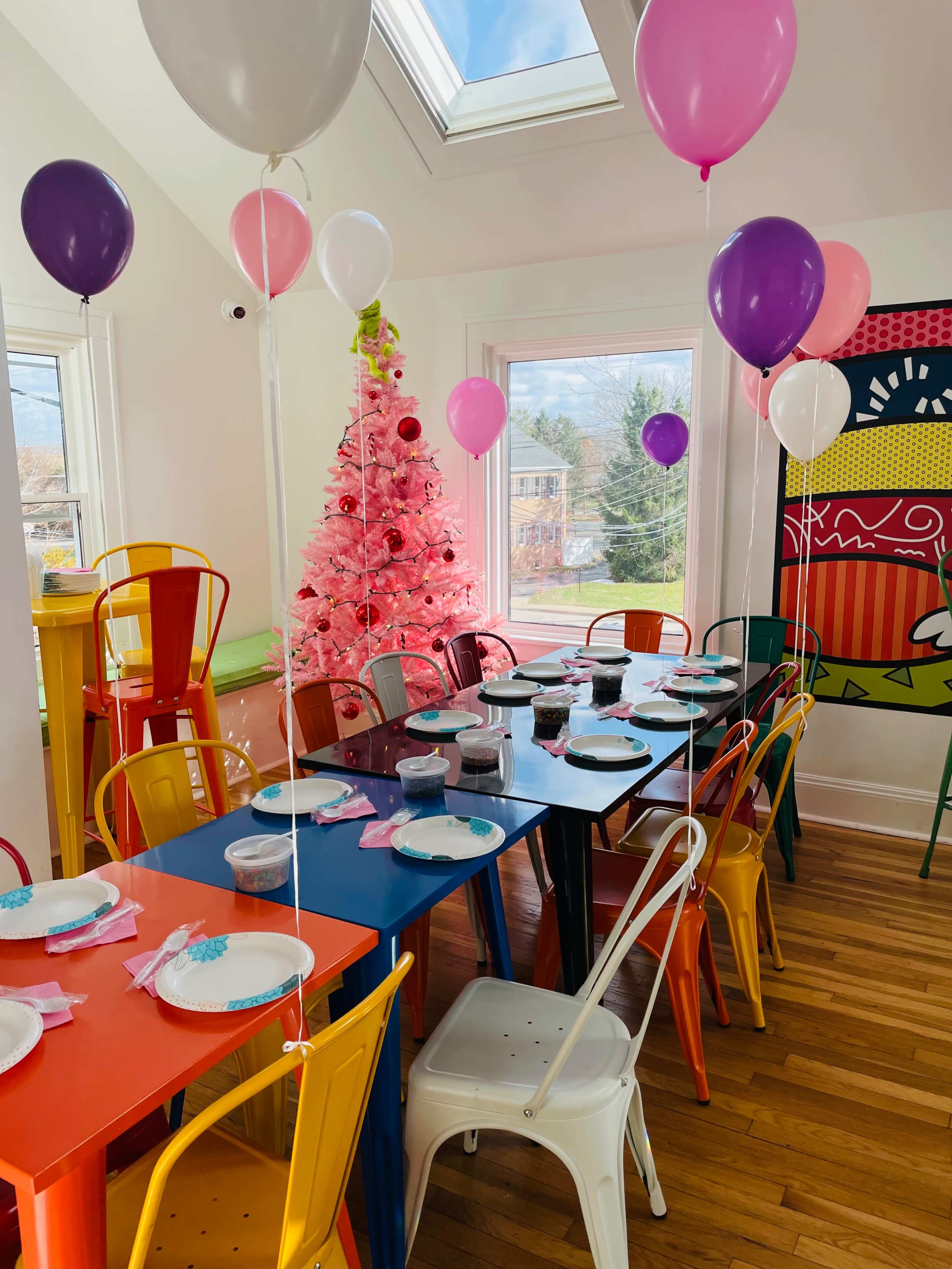 A colorful dining area is set for a celebration, featuring multiple tables with plates and drinks, decorated with balloons and a pink Christmas tree in the background.