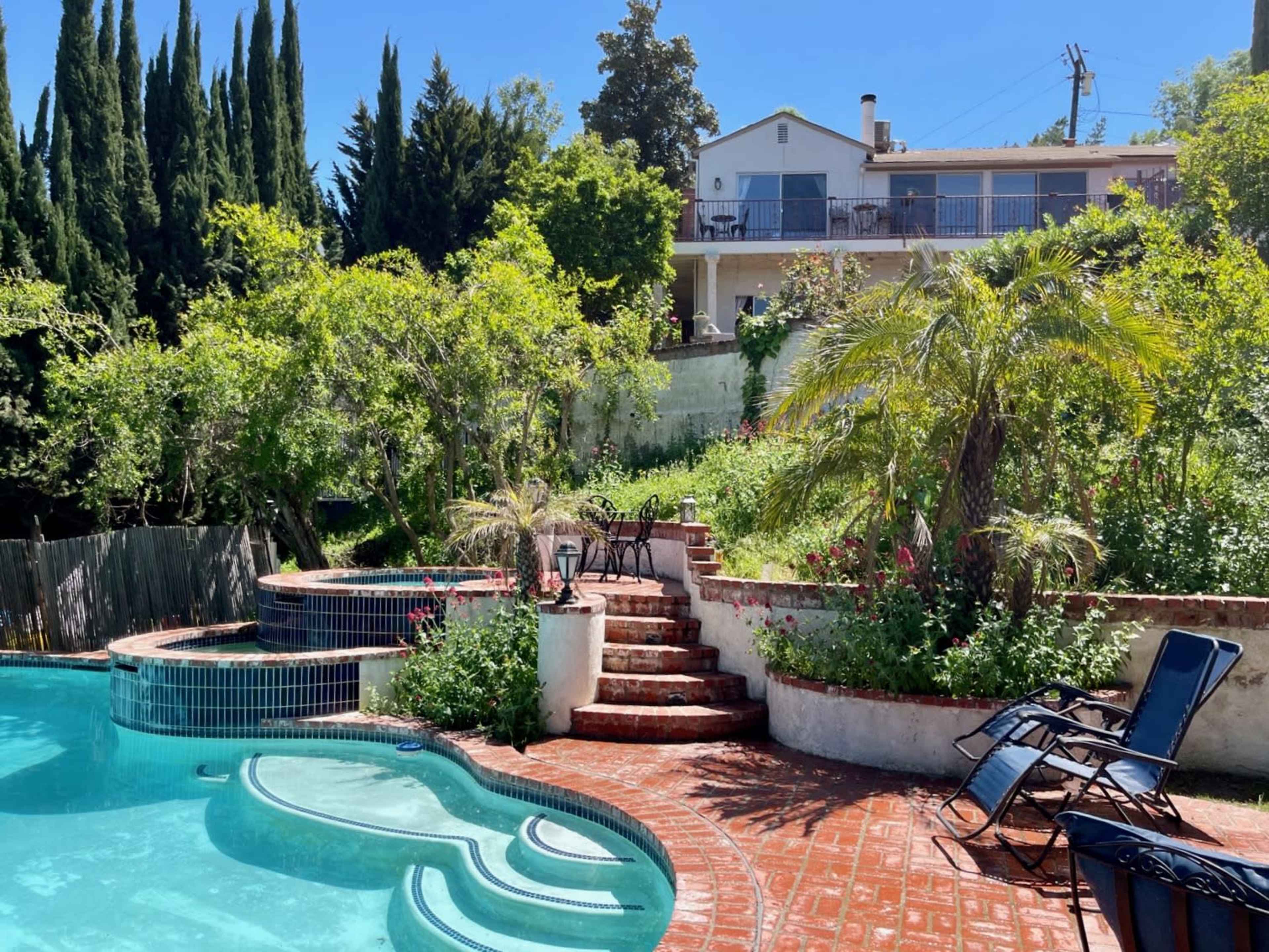 A tiered backyard features a swimming pool with a curved edge, steps leading to a garden area, and a two-story house in the background.