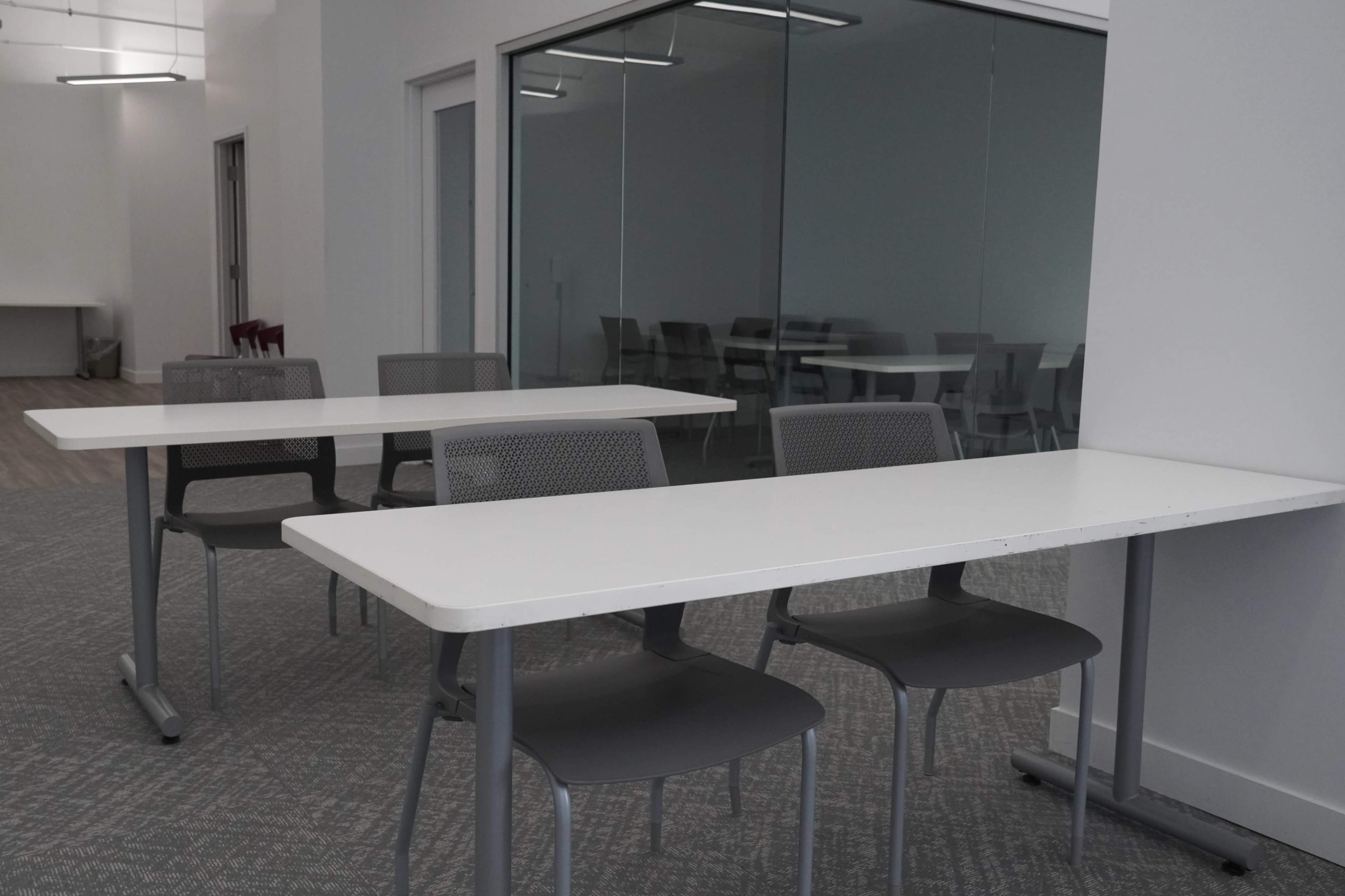 Two white tables with gray chairs in a bright, modern workspace, with additional chairs visible through a glass wall in the background.