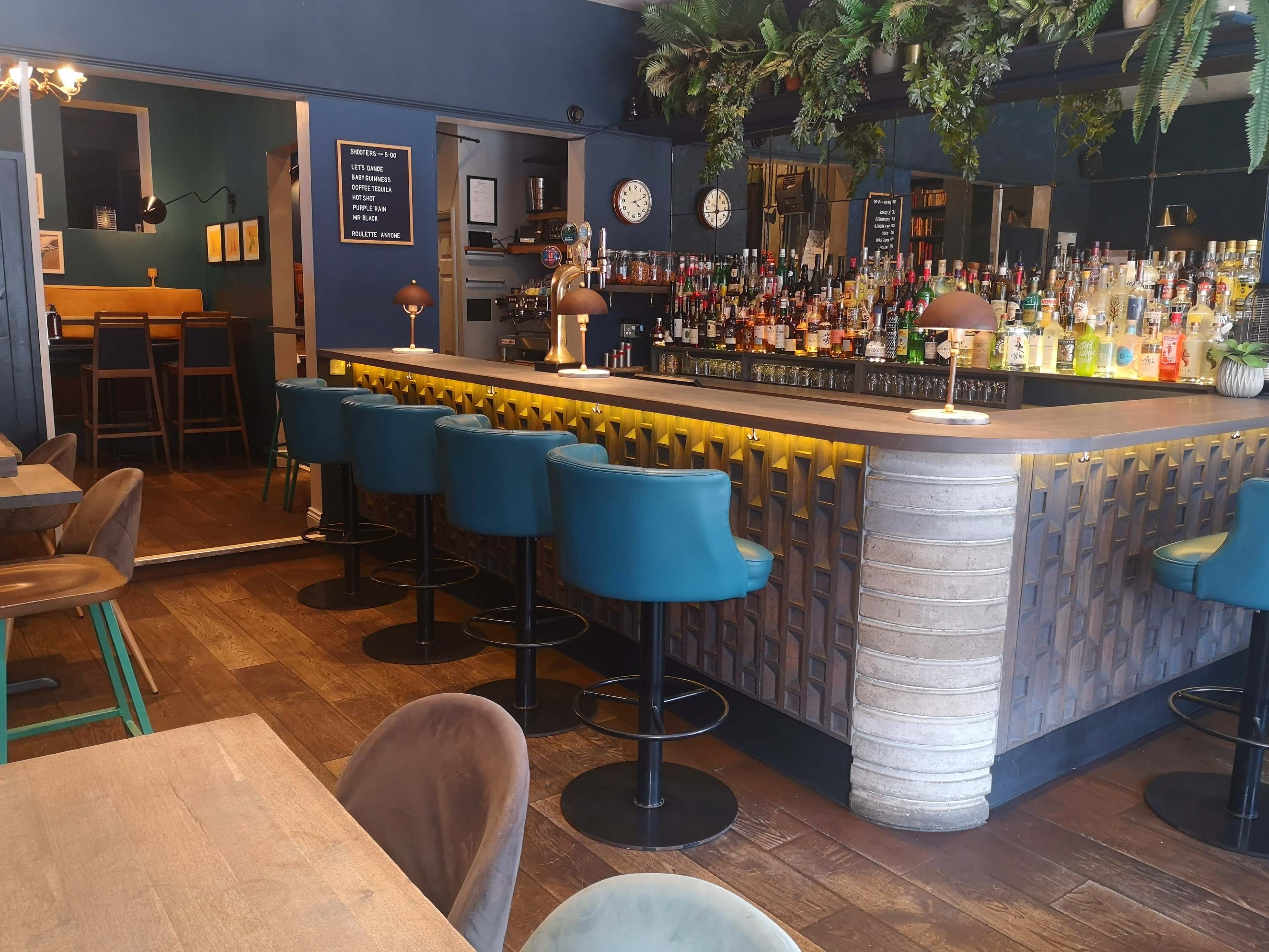 The image shows a modern bar area with teal-colored stools, a well-stocked backlit bar, and wooden flooring, adjacent to a dining space.