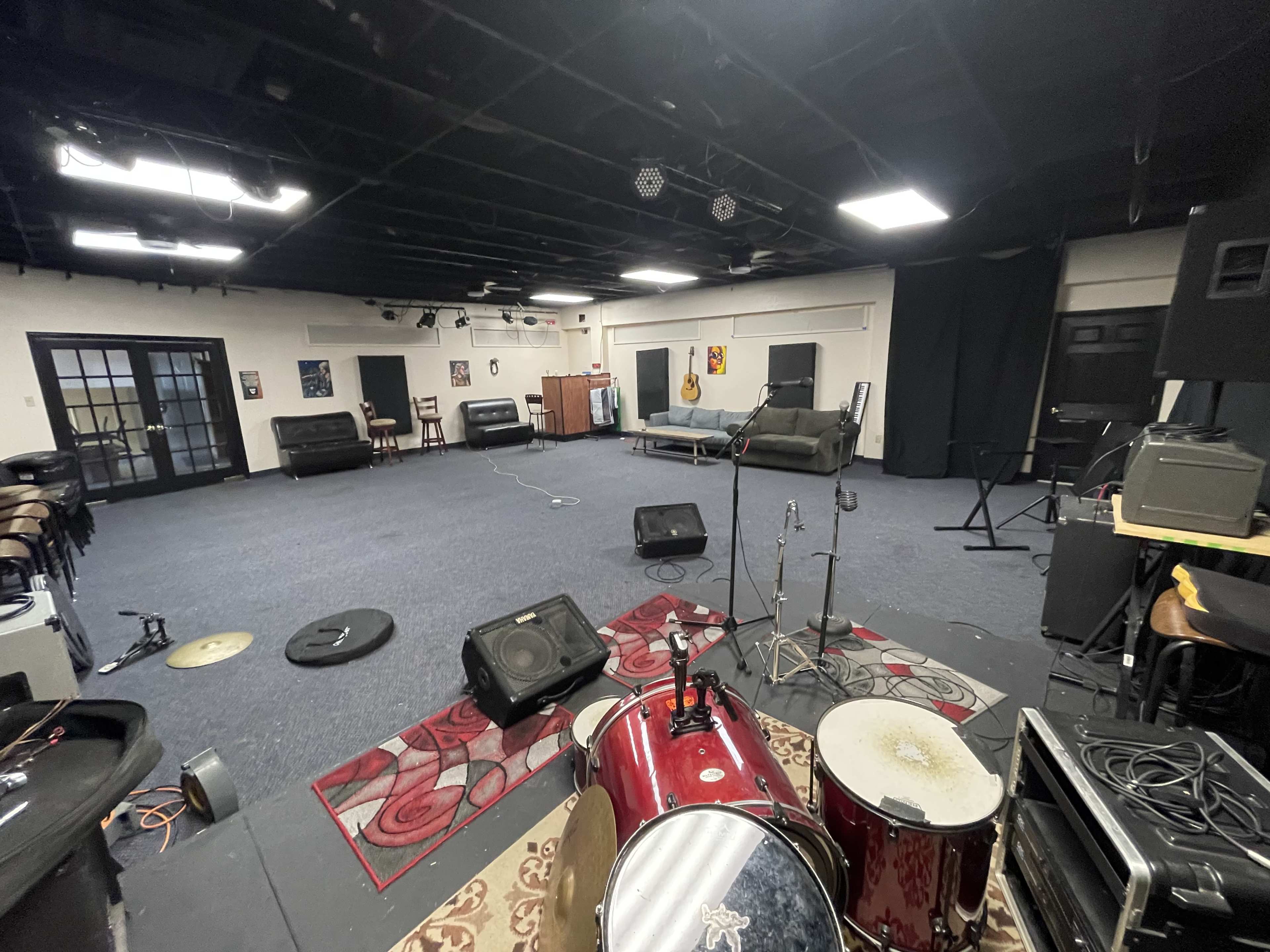 The image shows a spacious rehearsal studio with musical instruments, amplifiers, and seating arranged around a carpeted area.