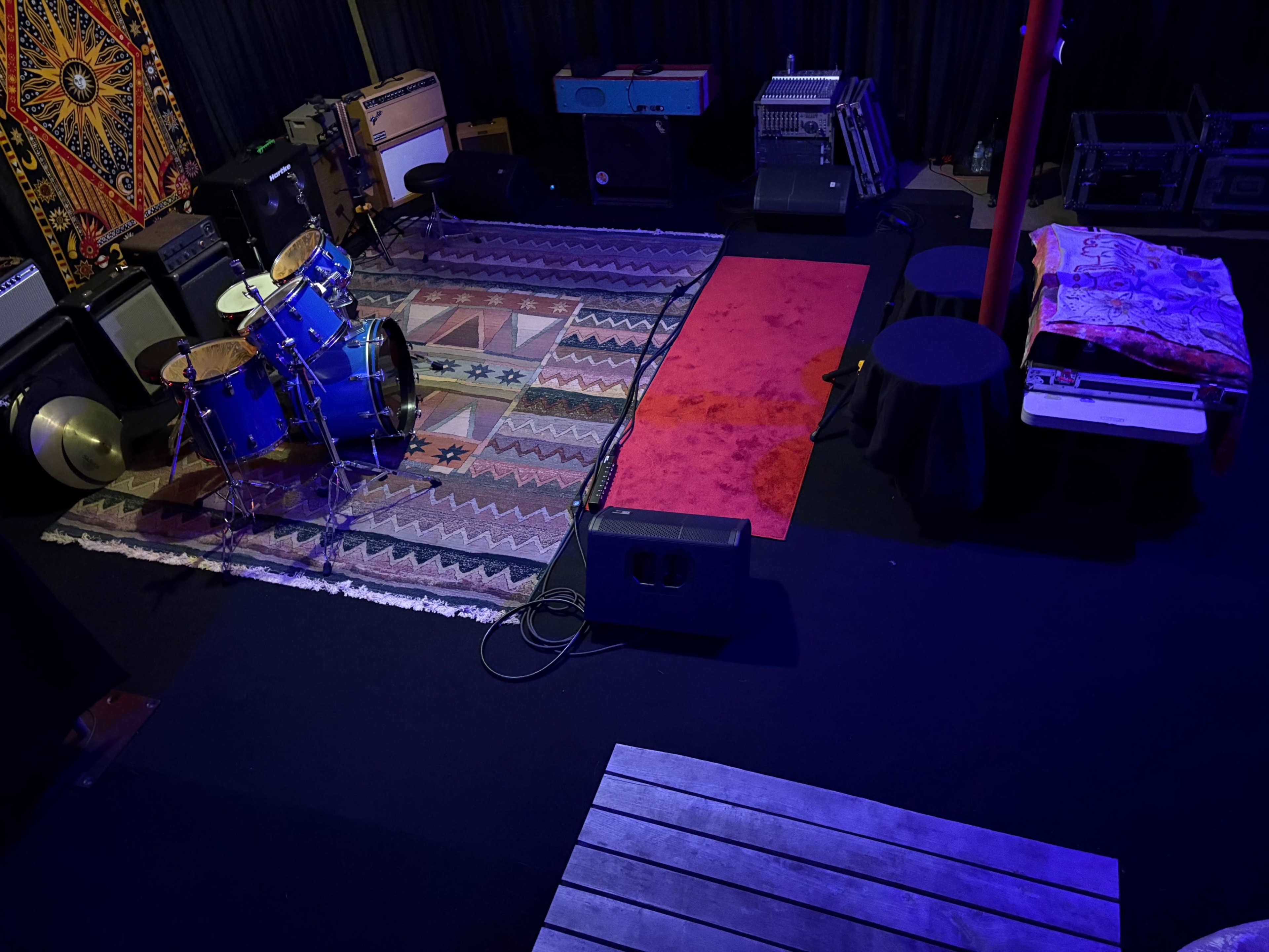The image shows a music rehearsal space featuring a drum set on a patterned rug, amplifiers in the background, and various equipment arranged around a red carpeted area.