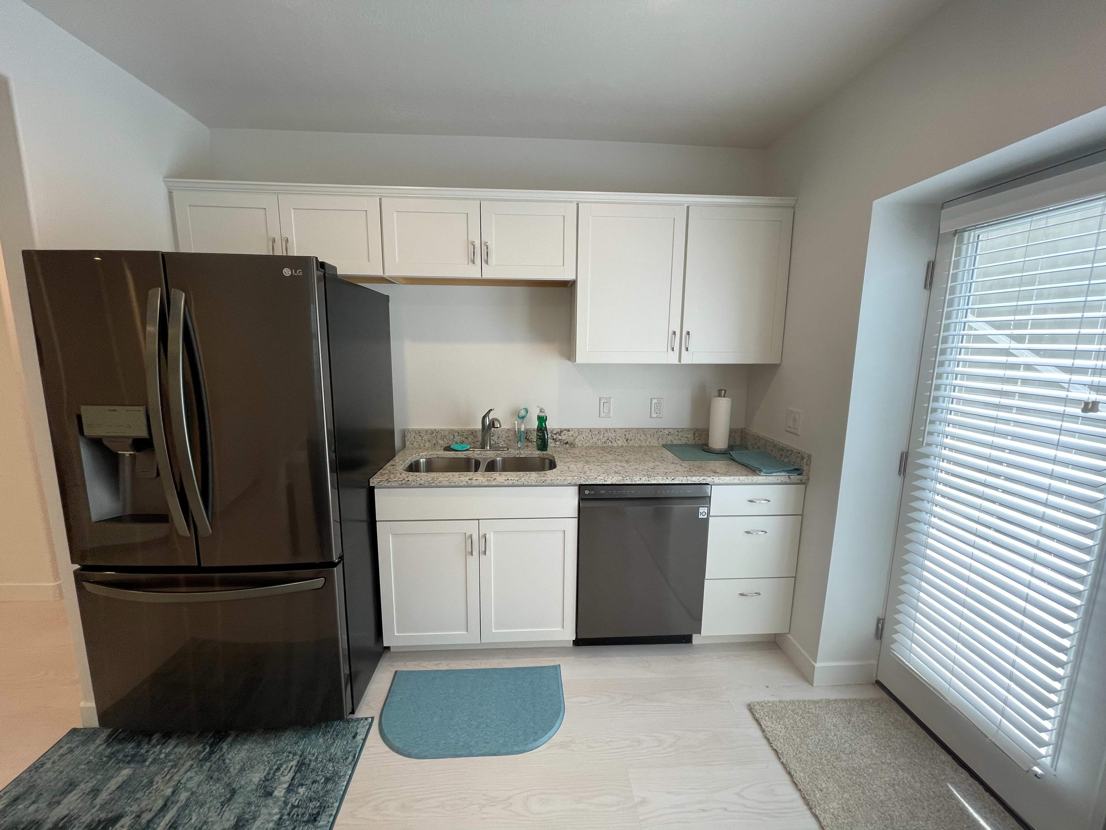 A modern kitchen with white cabinets, a granite countertop, a black refrigerator, a stainless steel dishwasher, and a window with blinds.