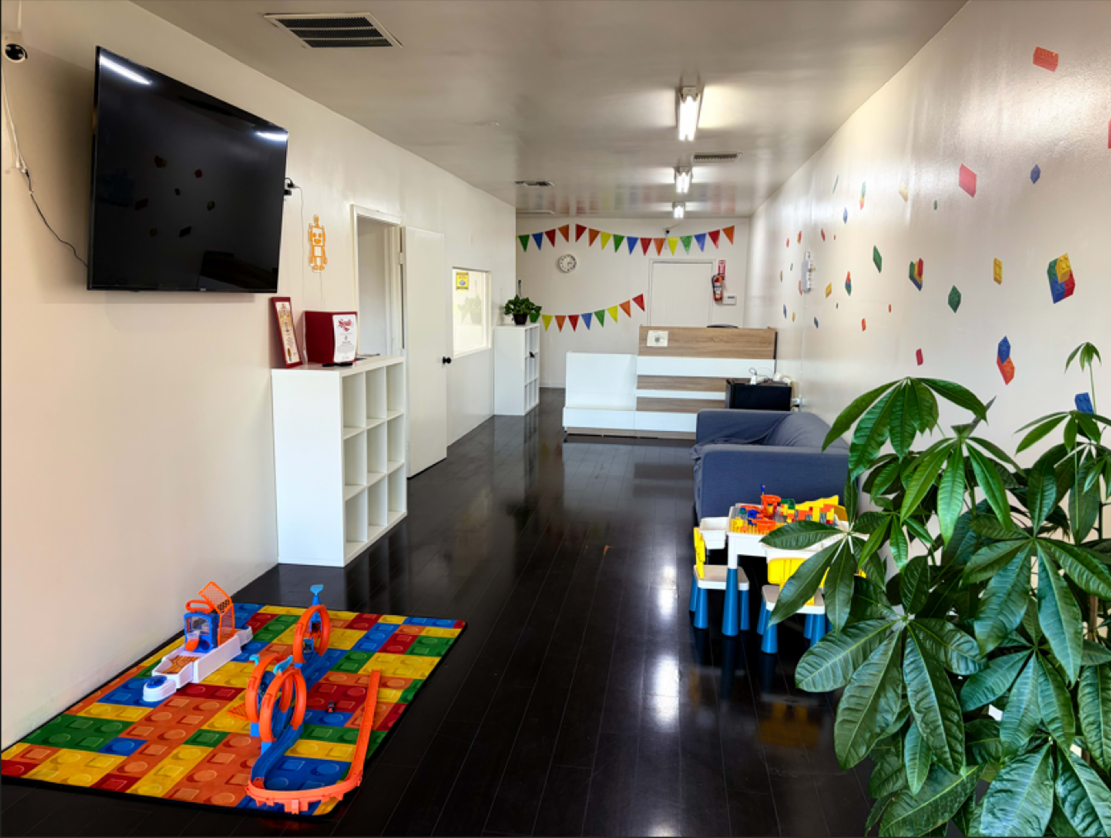 The image shows a bright, minimalist room with a play area featuring colorful mats and toys, along with seating and decorative elements like banners on the walls.