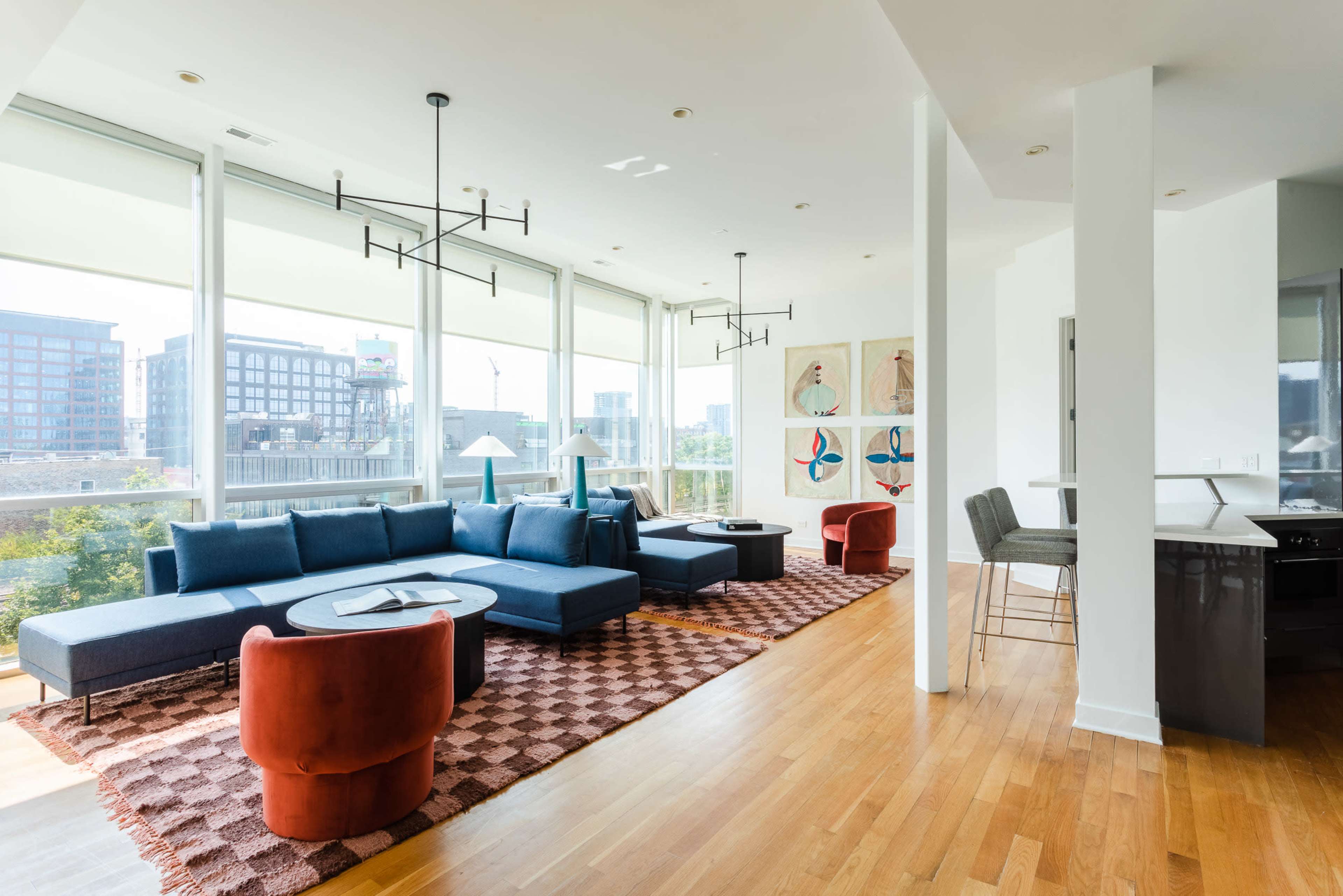 A modern living space featuring a blue sectional sofa, two red accent chairs, and a large area rug, with large windows offering a view of the city.