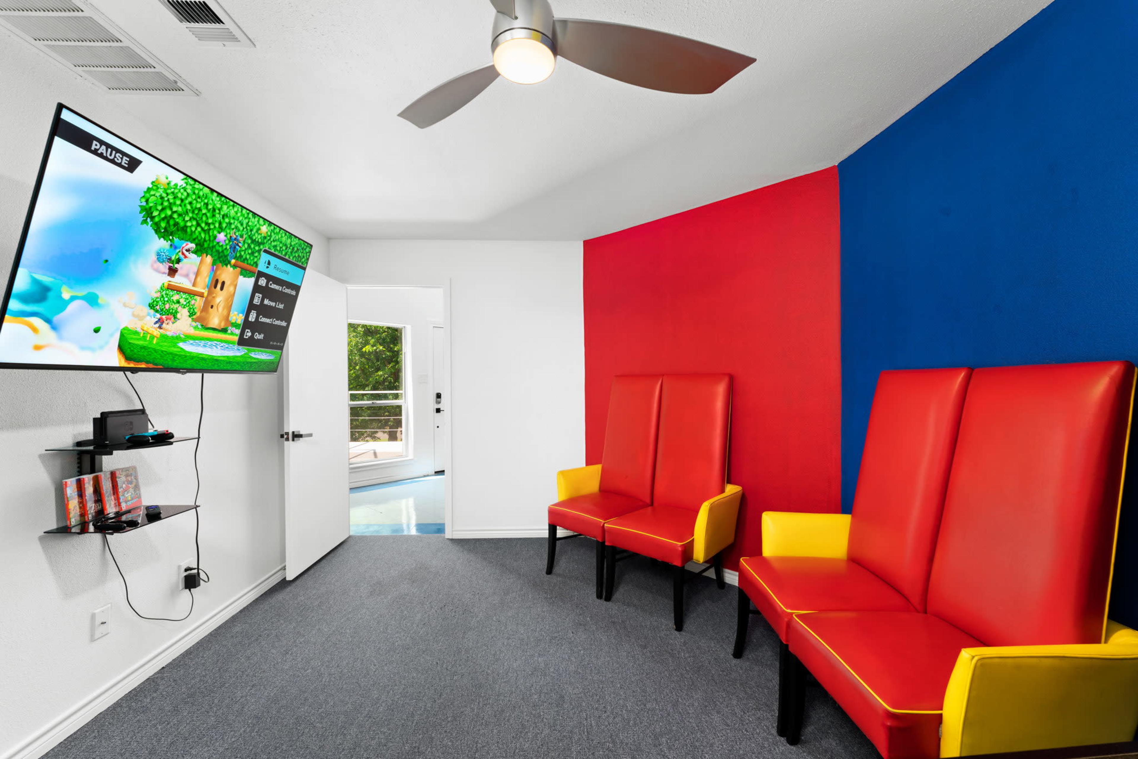 The room features two red and yellow chairs facing a wall-mounted TV displaying a video game menu, with a blue accent wall and an entry door leading outside.