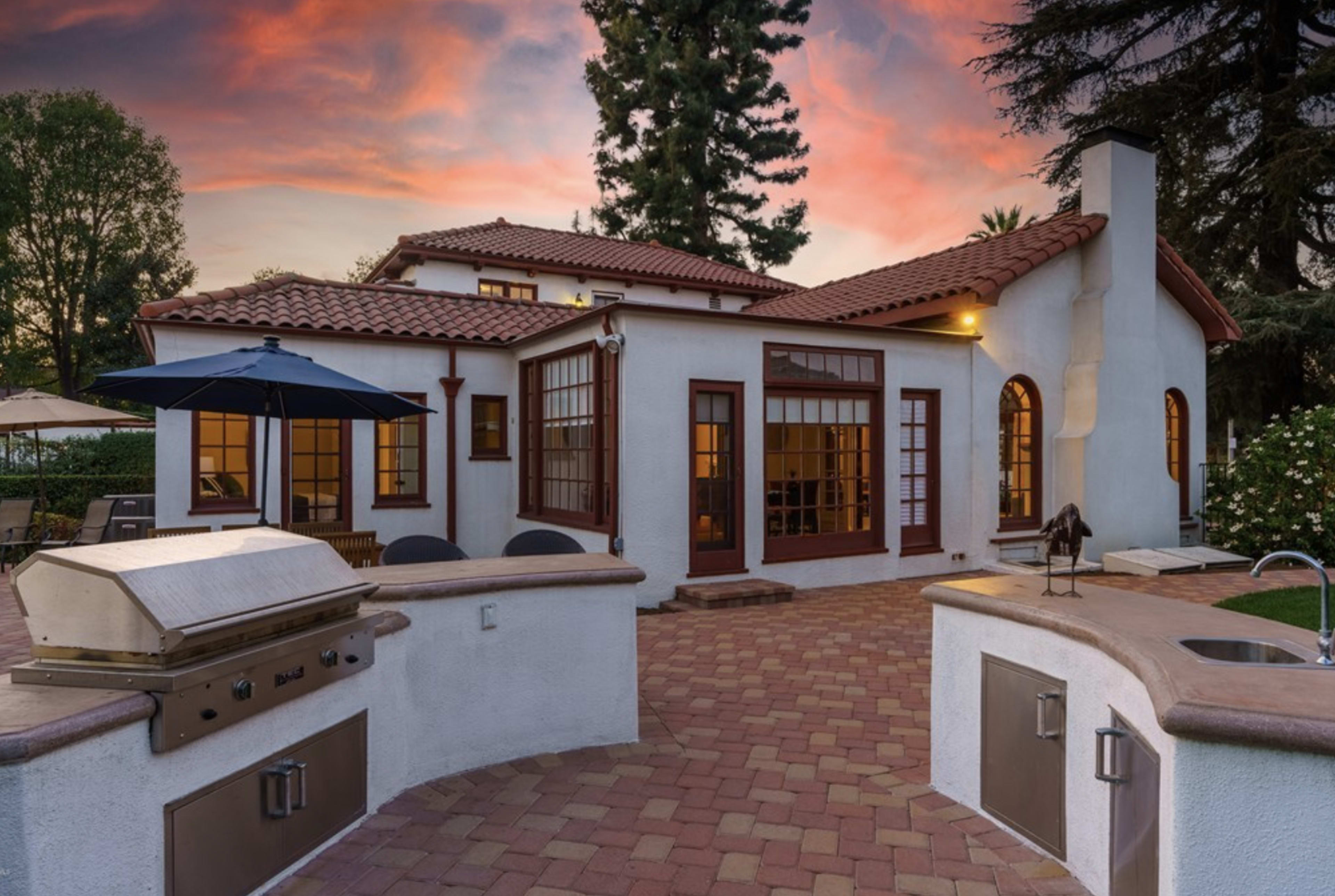 A landscaped backyard with a patio featuring a built-in grill and outdoor seating, adjacent to a house with large windows and a tiled roof under a colorful sunset sky.