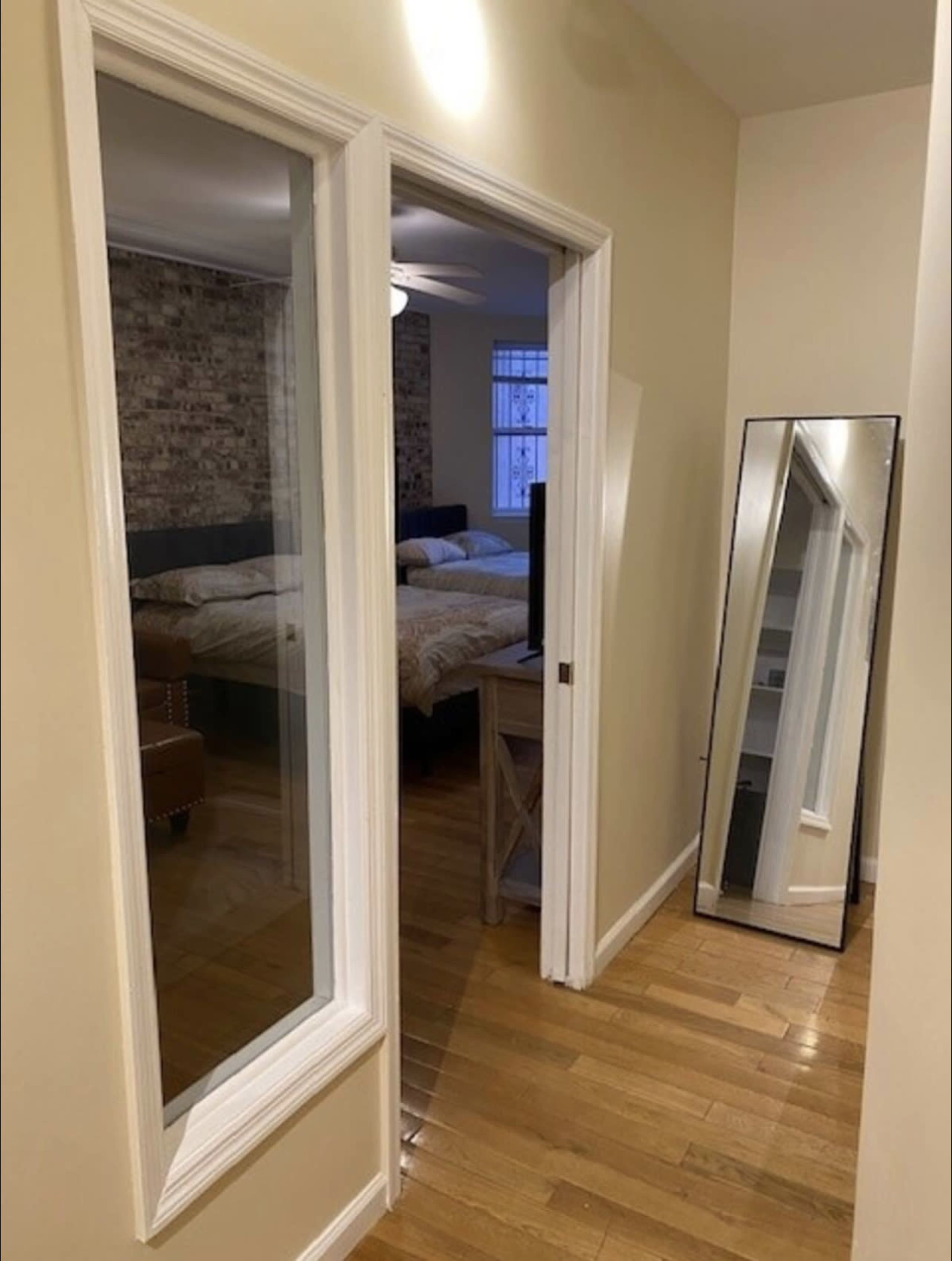A narrow hallway features a doorway leading to a bedroom, with a large mirror on one side and a window in the background.