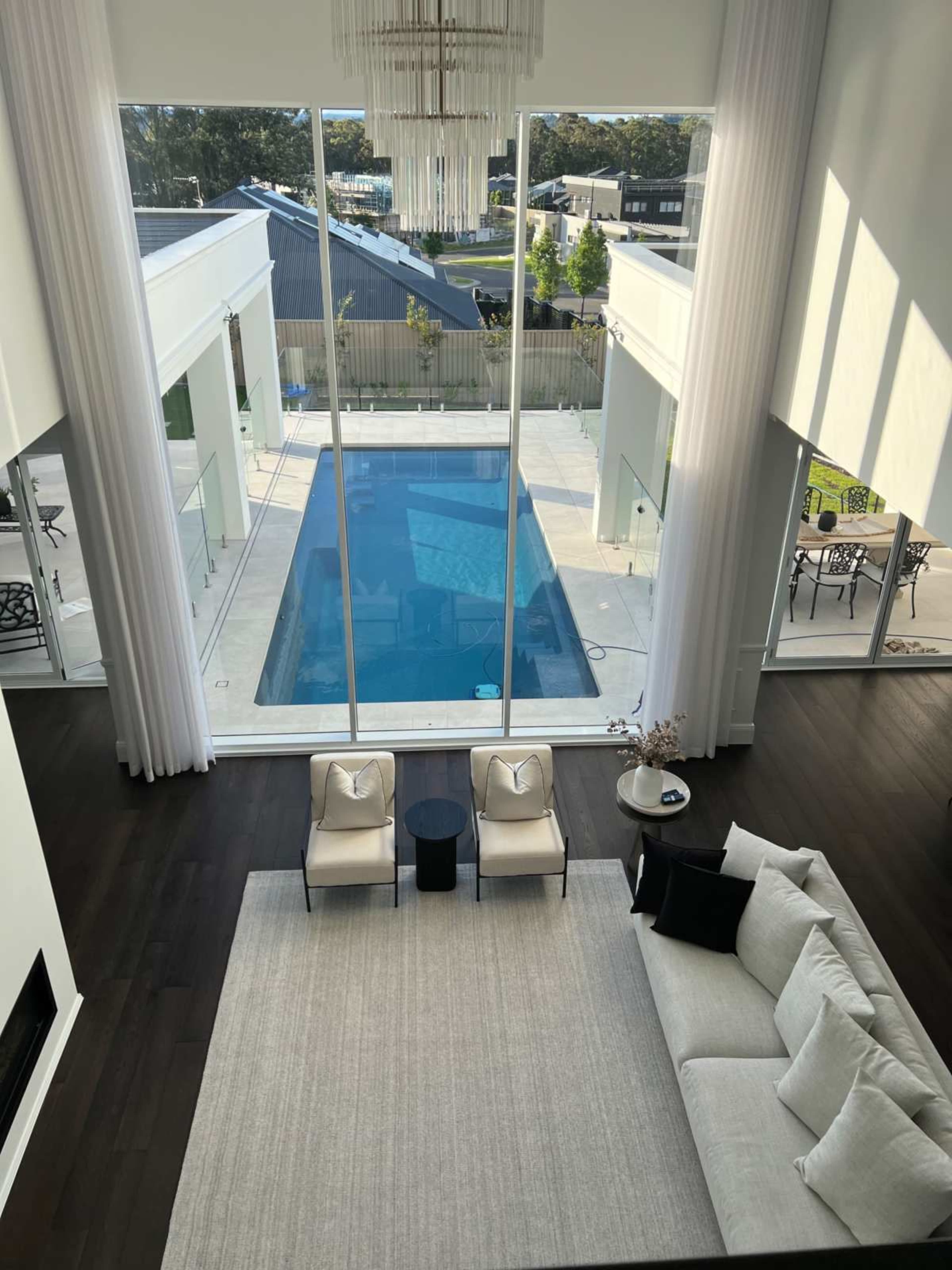 The image shows a modern living room with large windows overlooking a pool area and outdoor seating, featuring a light-colored sofa, two chairs, and a coffee table on a rug.