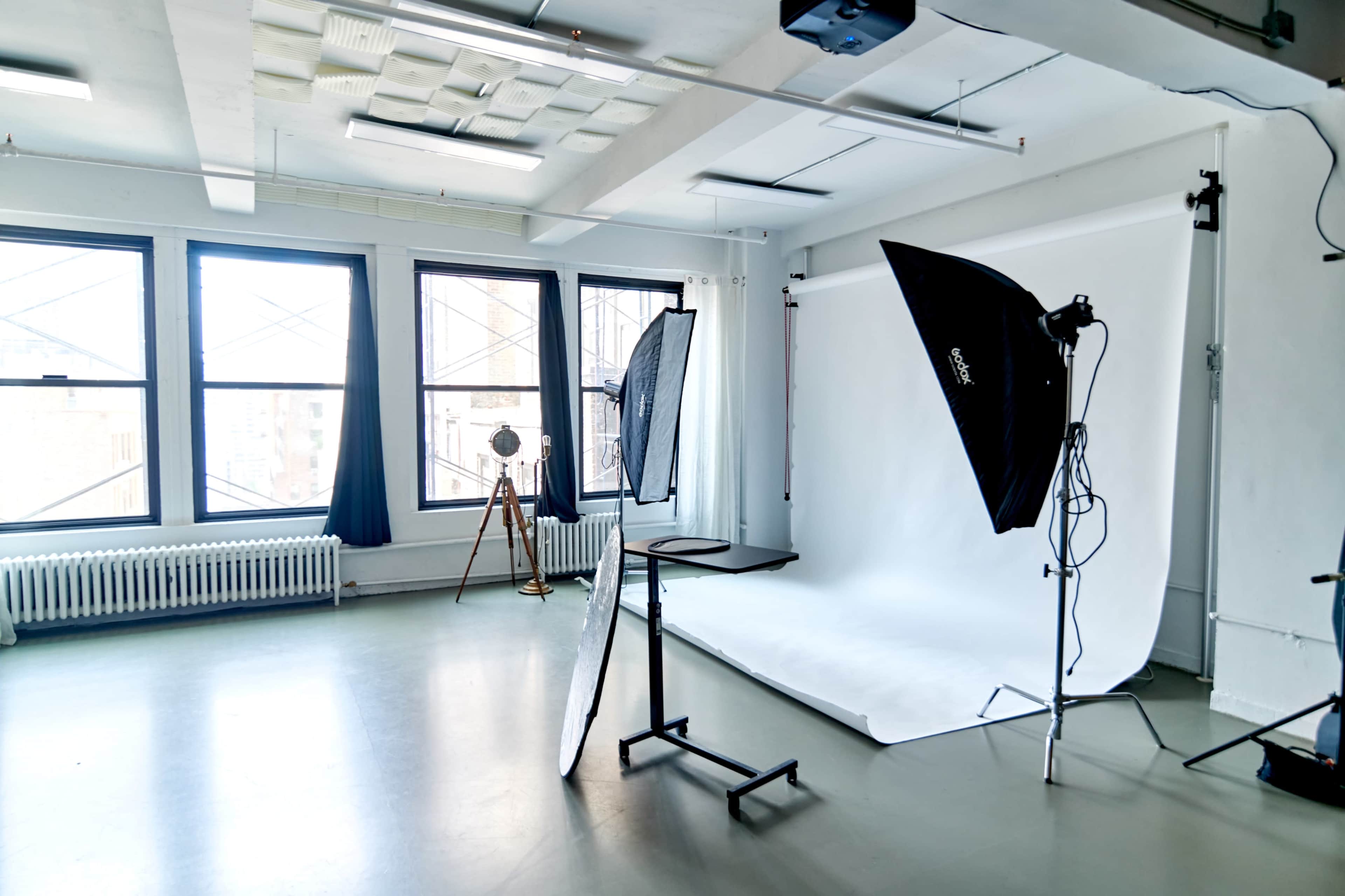A photography studio equipped with a white backdrop, lighting equipment, and large windows providing natural light.