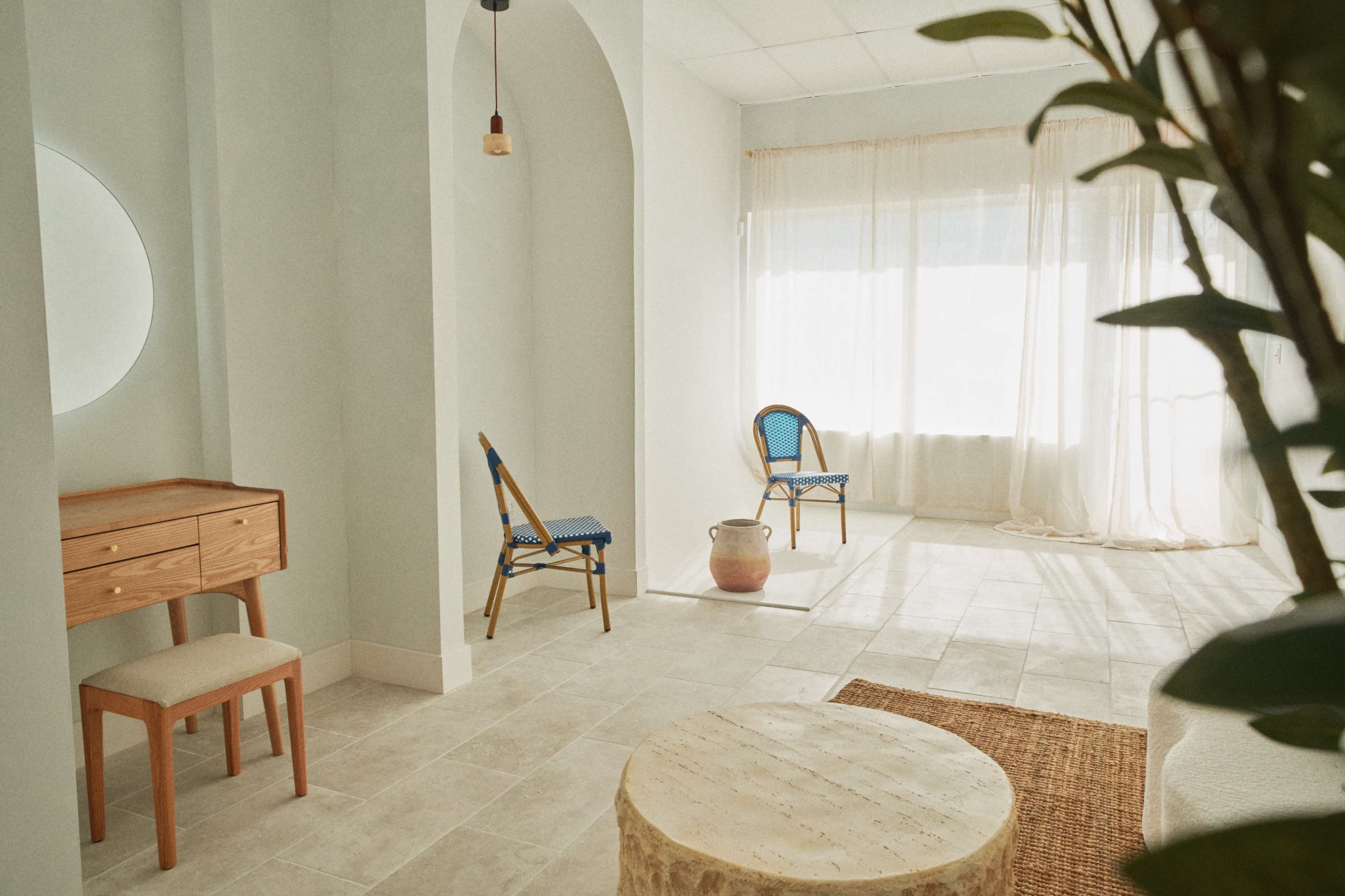 The image shows a minimalistic interior room featuring light-colored walls, a circular mirror, two chairs with woven backs, a small wooden table, and a potted plant.