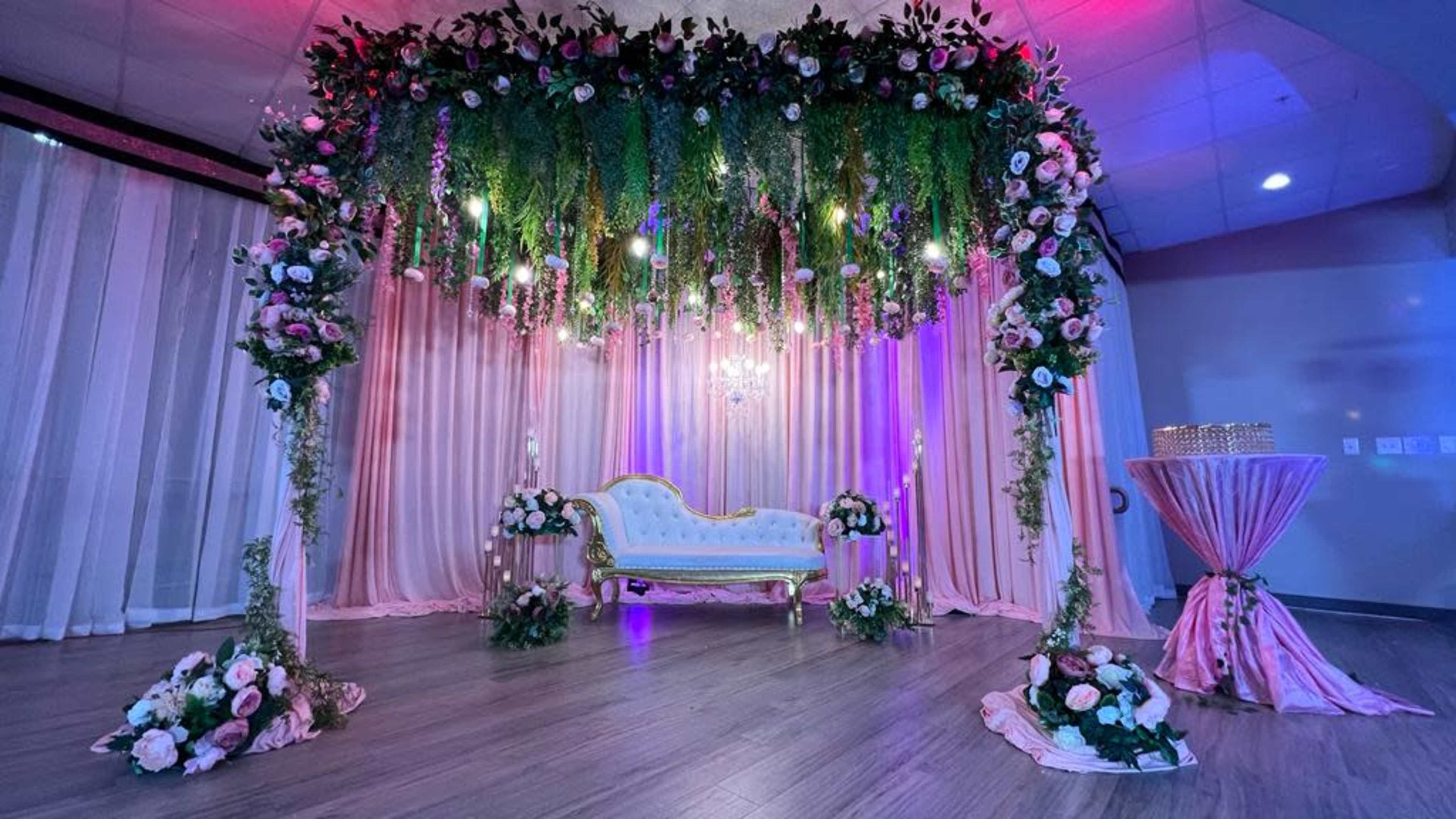 The scene features a beautifully decorated event space with a lavish floral backdrop, a vintage white sofa under hanging greenery and lights, and a pink draped table nearby.