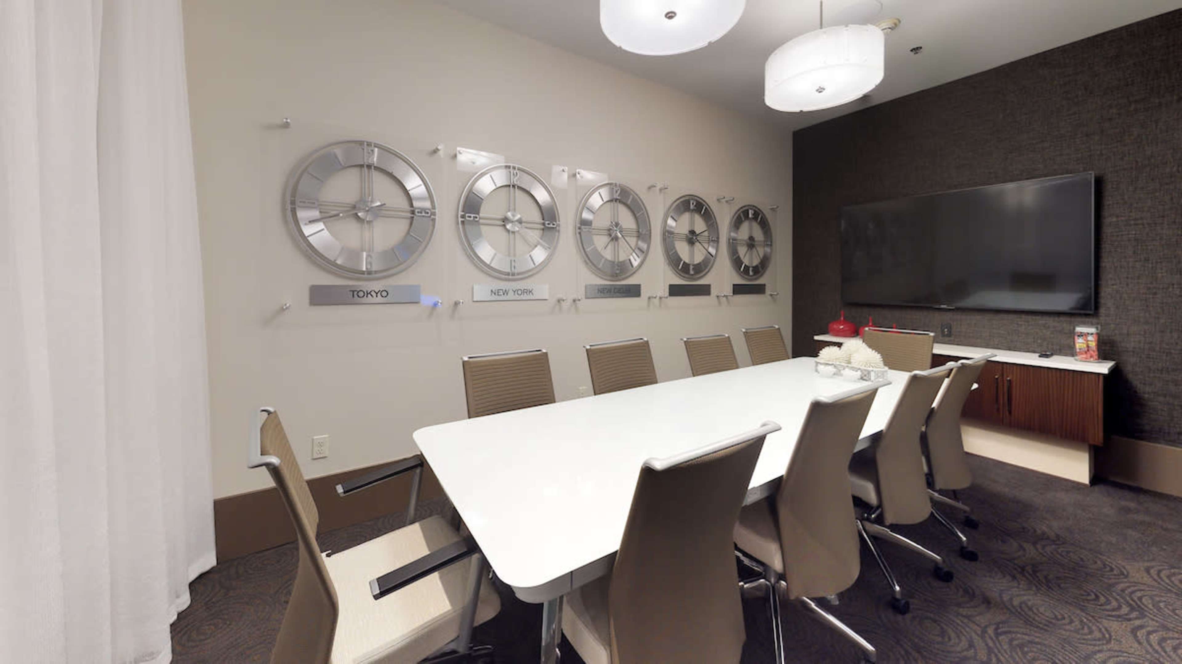 A modern conference room features a long white table surrounded by beige chairs, with wall clocks from different time zones displayed on the wall.