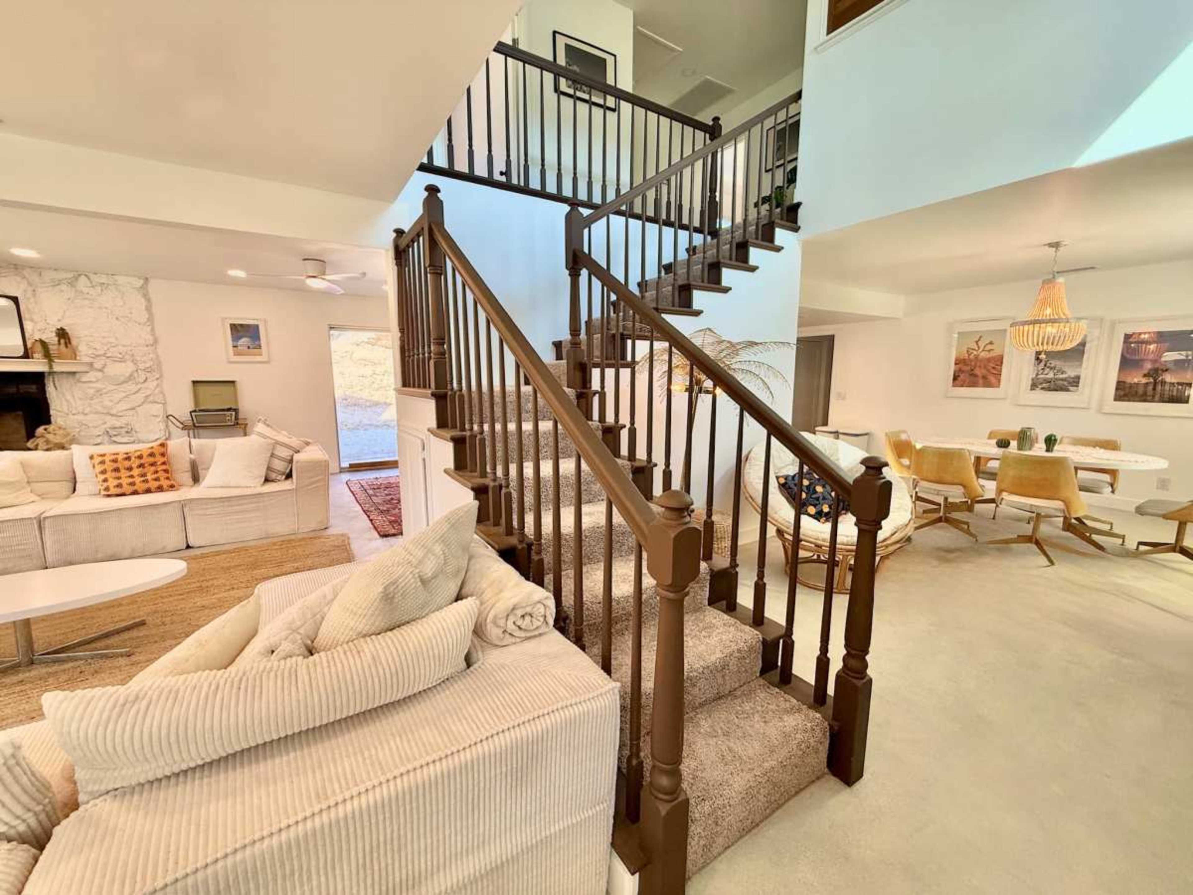 The image shows a modern interior of a home featuring a staircase, a living area with a beige sofa, and a dining area with chairs and a table.