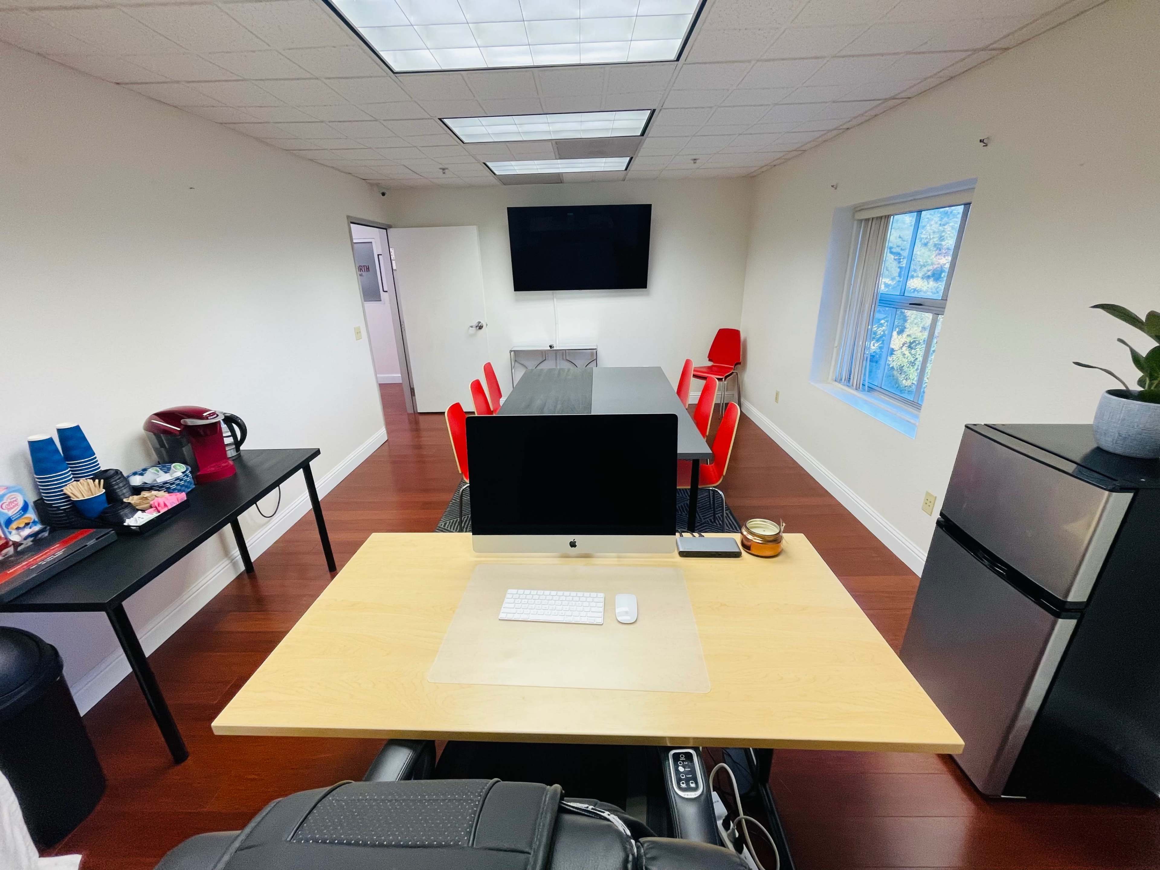 A modern office meeting room features a large conference table, a desktop computer on a wooden desk, and red chairs along with a kitchenette area.