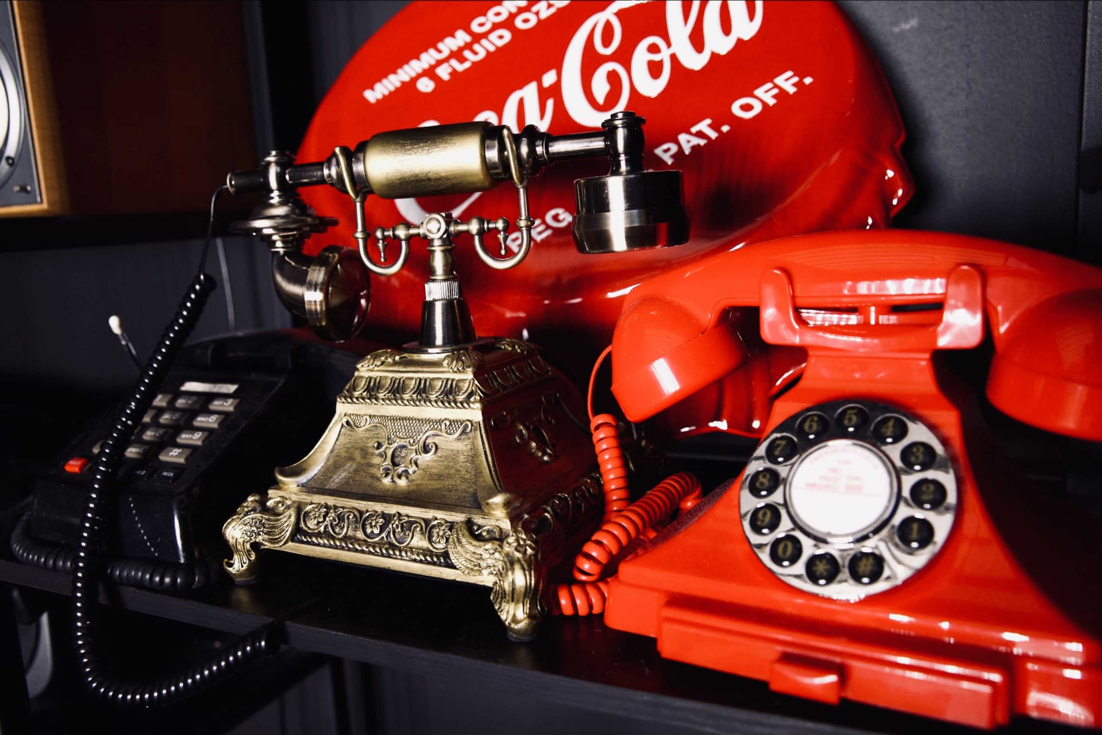 A vintage brass desk telephone alongside a bright red rotary phone, both positioned on a shelf near a Coca-Cola sign.