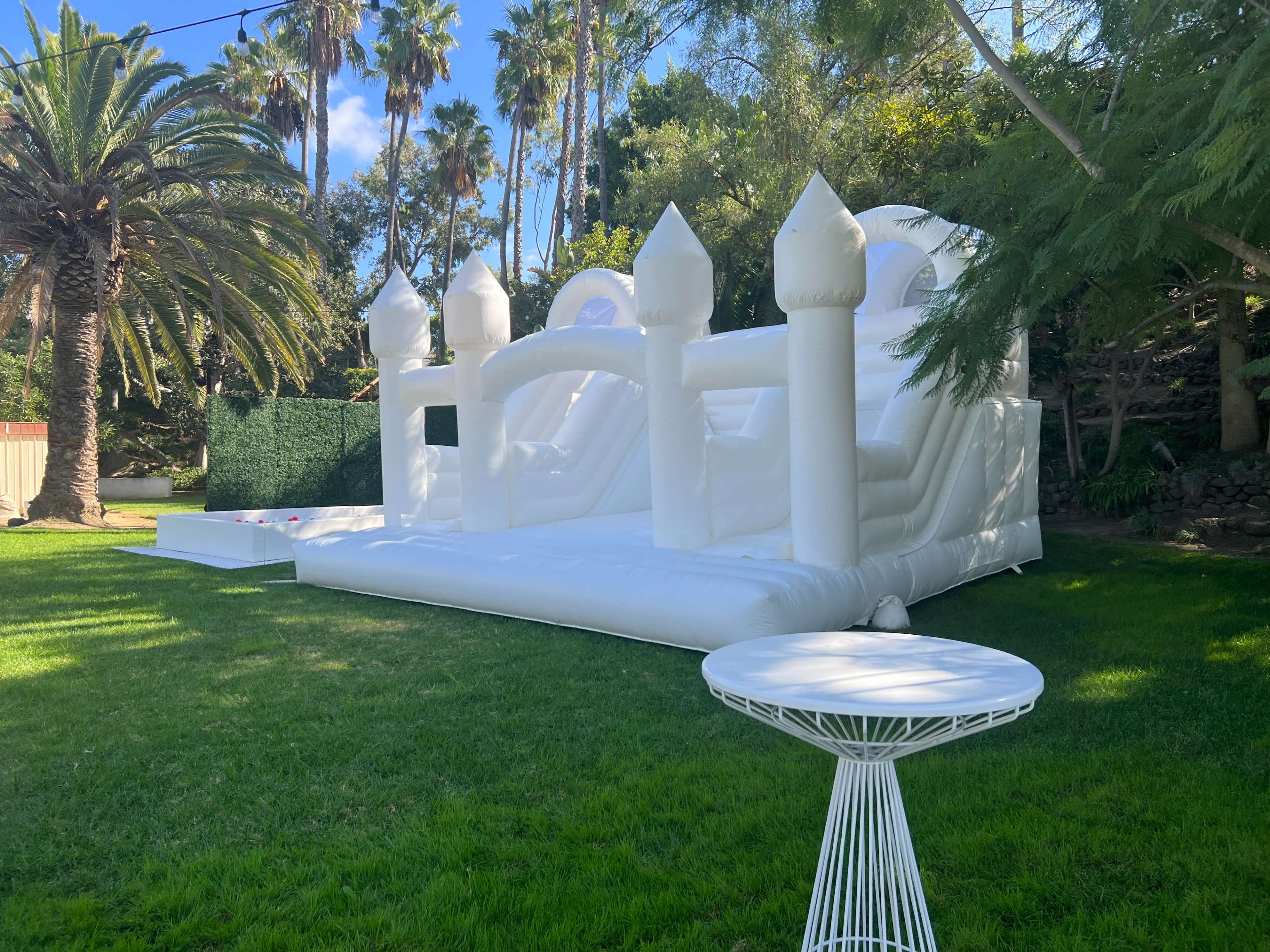 A large, white inflatable bounce house with turrets is set up on a grassy lawn surrounded by palm trees.