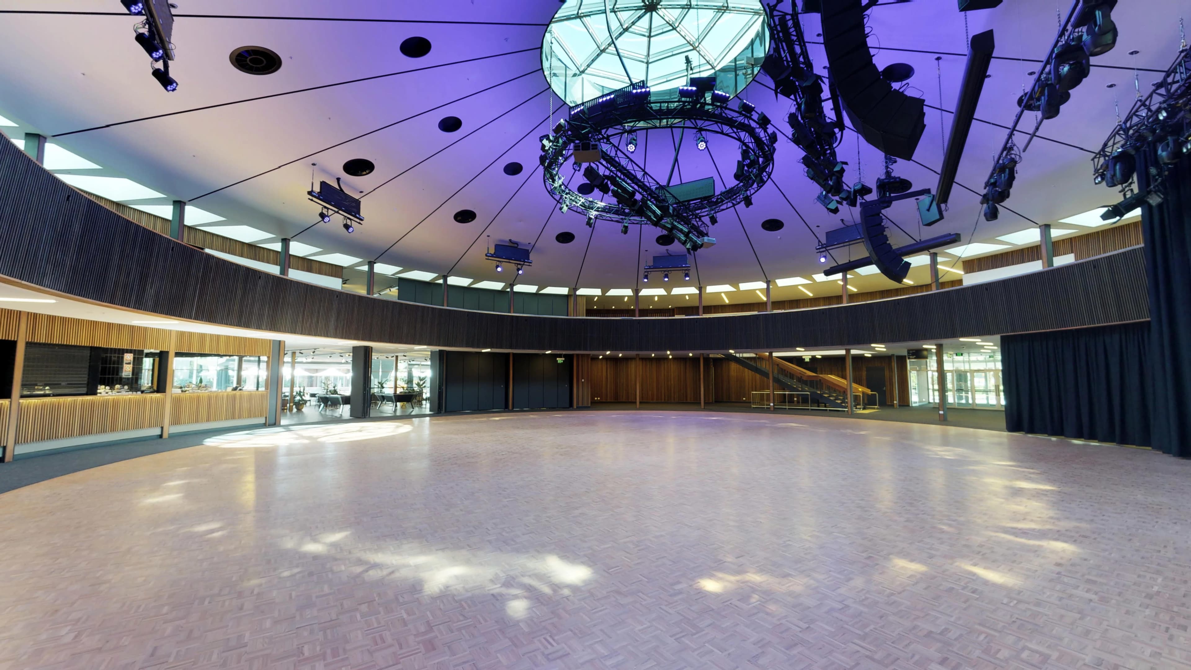 A spacious circular event hall with a glossy wooden floor, large windows, and a modern lighting setup overhead.
