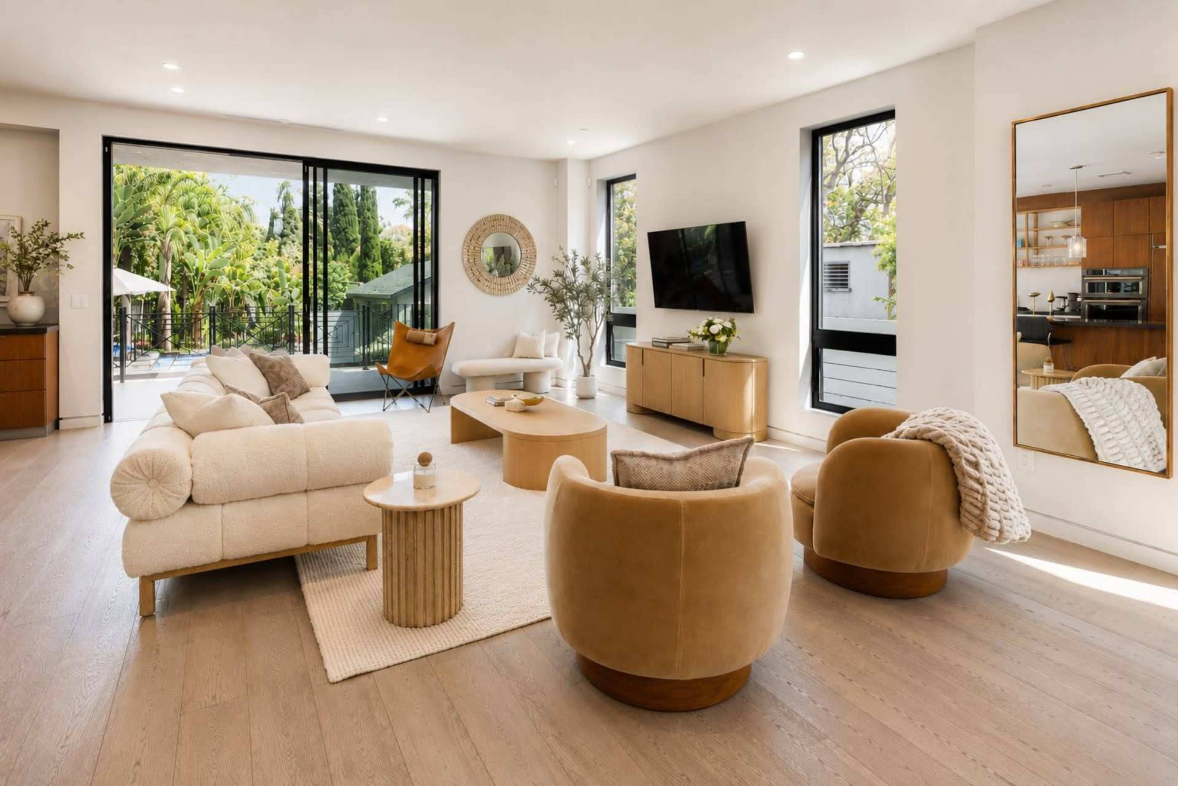 A modern living room features beige sofas, round chairs, and a coffee table, with large windows overlooking a pool area.