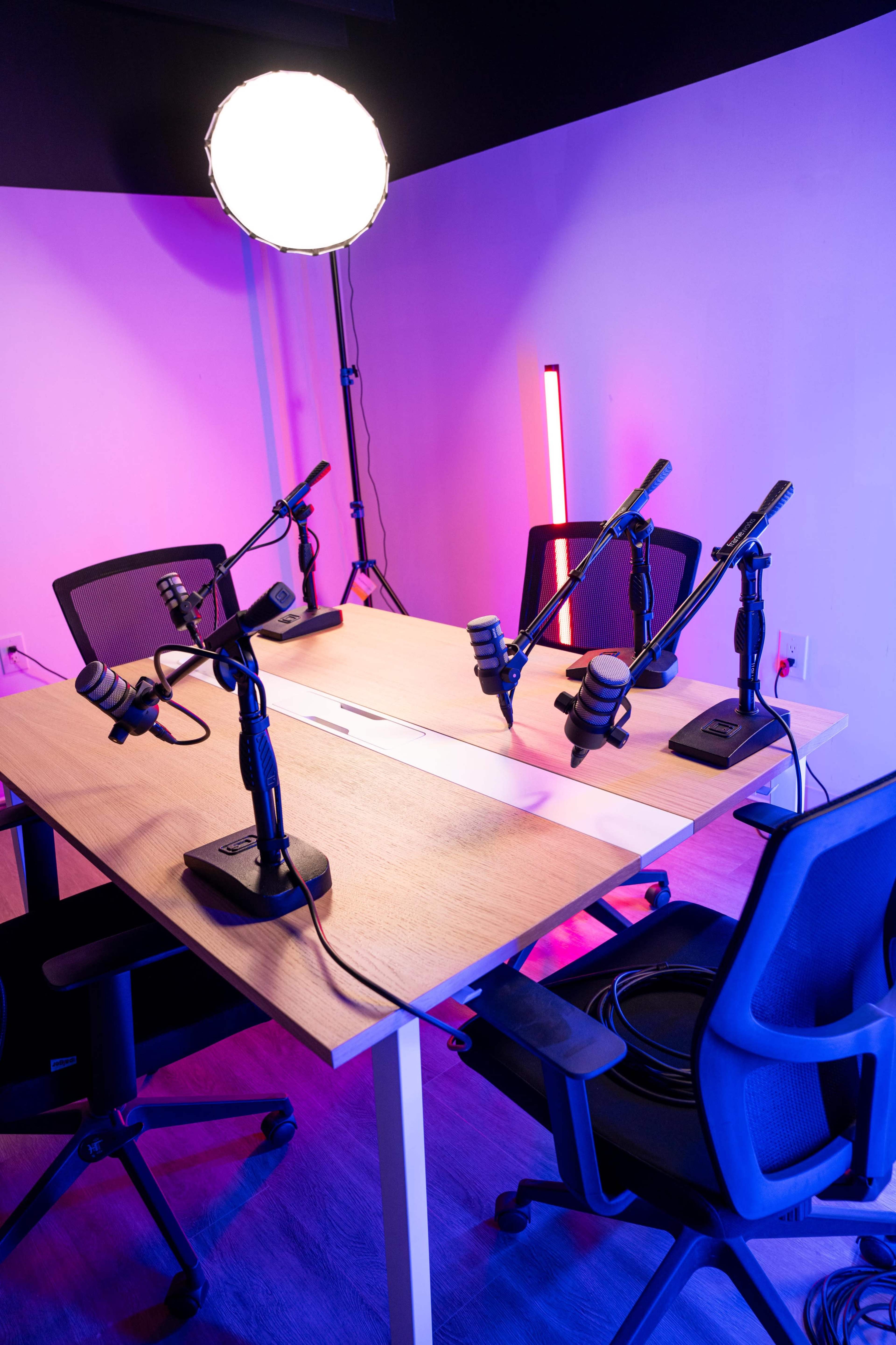 A podcast recording studio features a wooden table with four microphones on adjustable arms, two office chairs, and colorful lighting in the background.