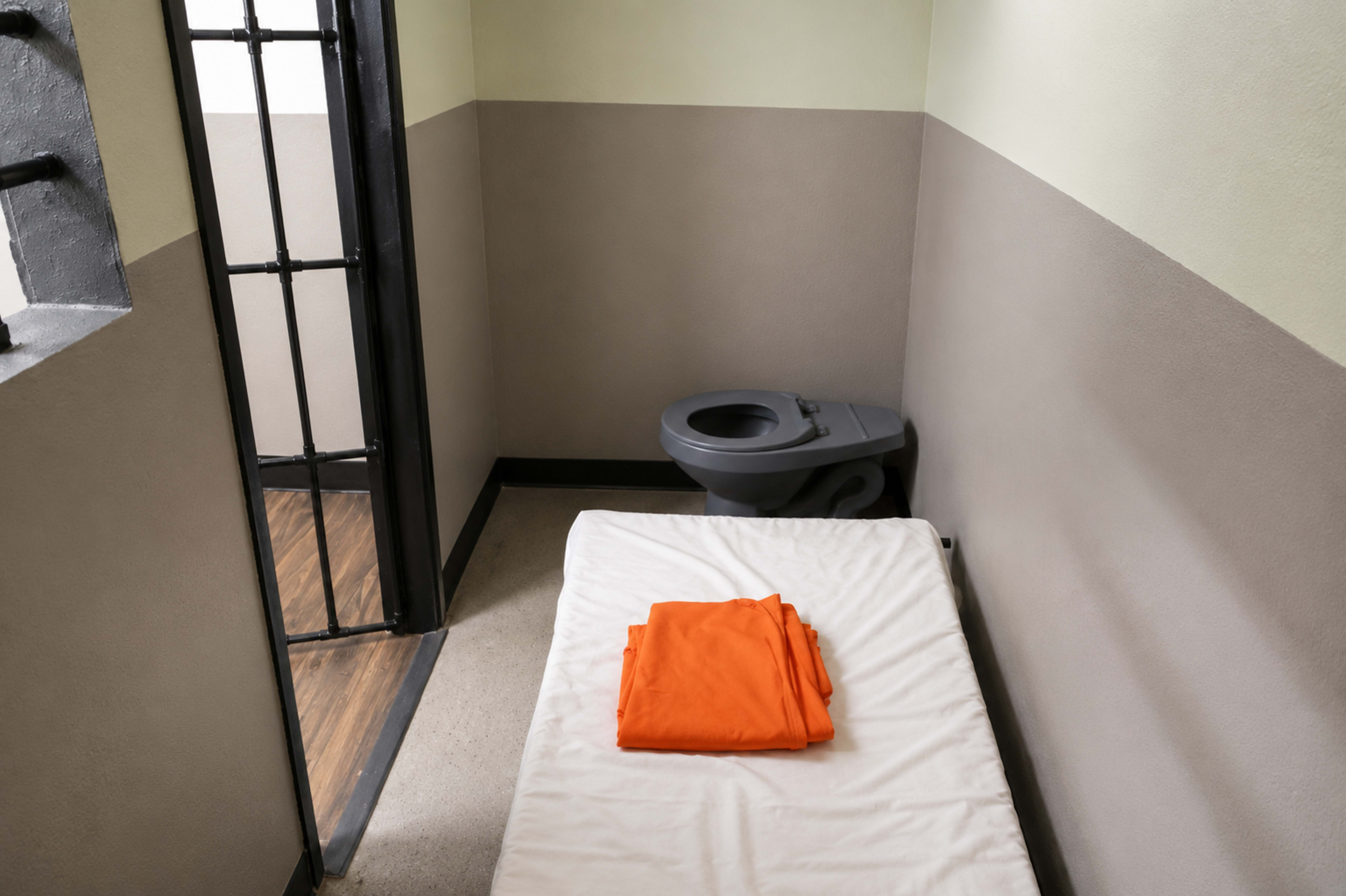The image shows a small, bare prison cell with a bed, an orange jumpsuit neatly folded on the mattress, a toilet, and a barred door.