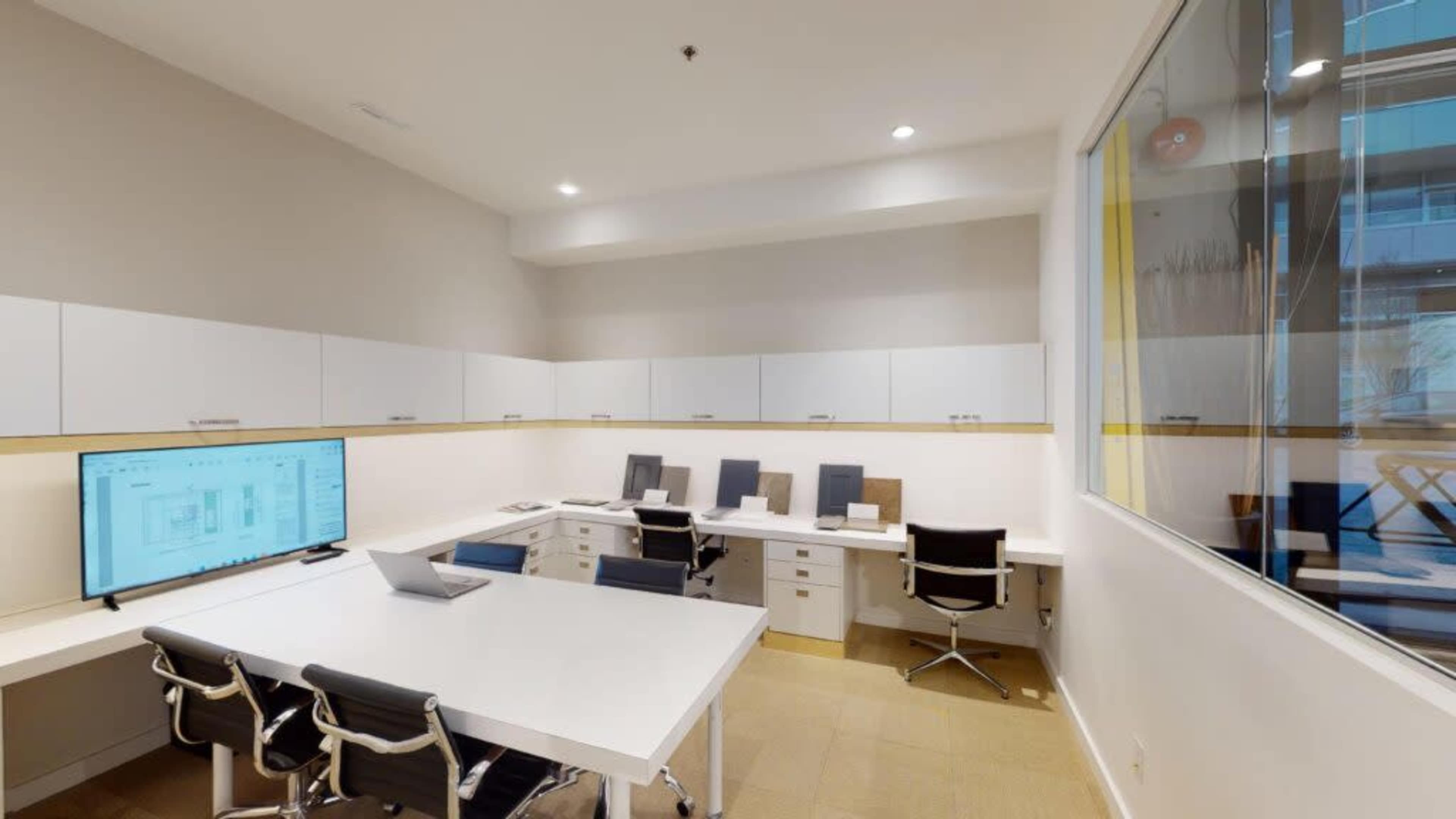 A modern office space featuring a white conference table, several black chairs, and a large screen displaying project layouts against a neutral wall.
