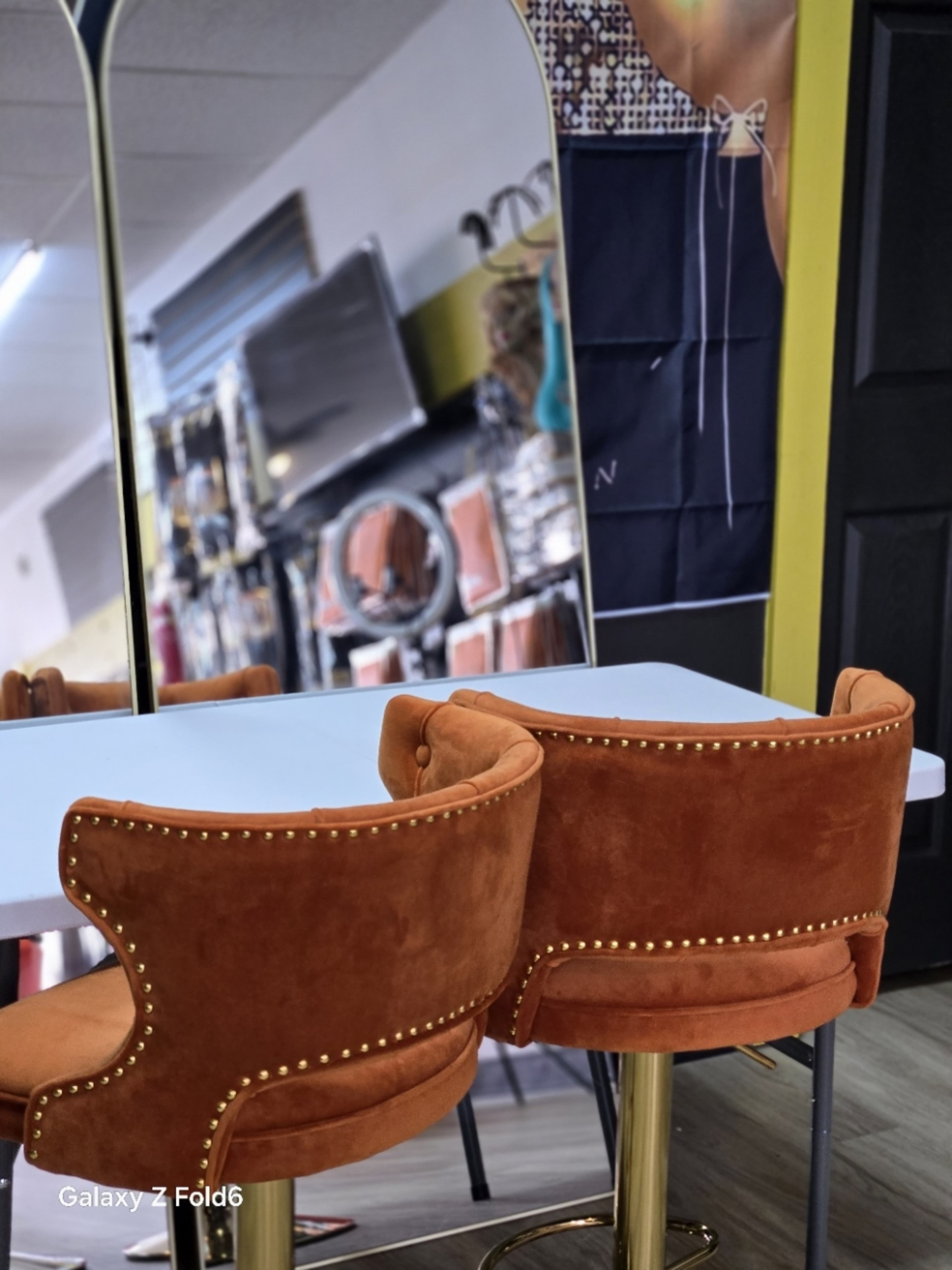 The image shows two orange velvet chairs with studded detailing positioned at a white table in a store setting that features mirrors and decorative items in the background.