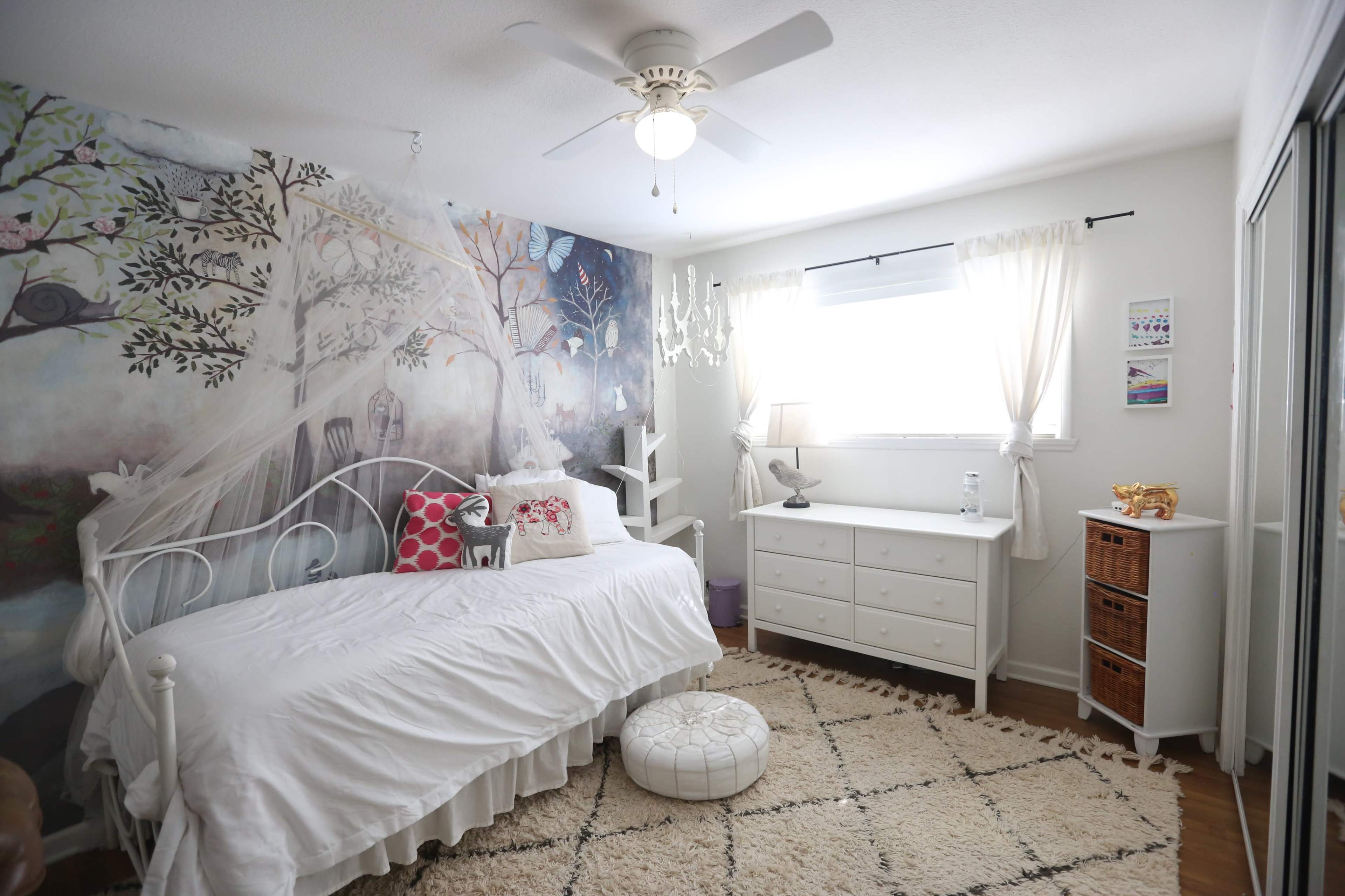 The image shows a bright bedroom with a white daybed, decorative pillows, a dresser, and a mural featuring trees on one wall.