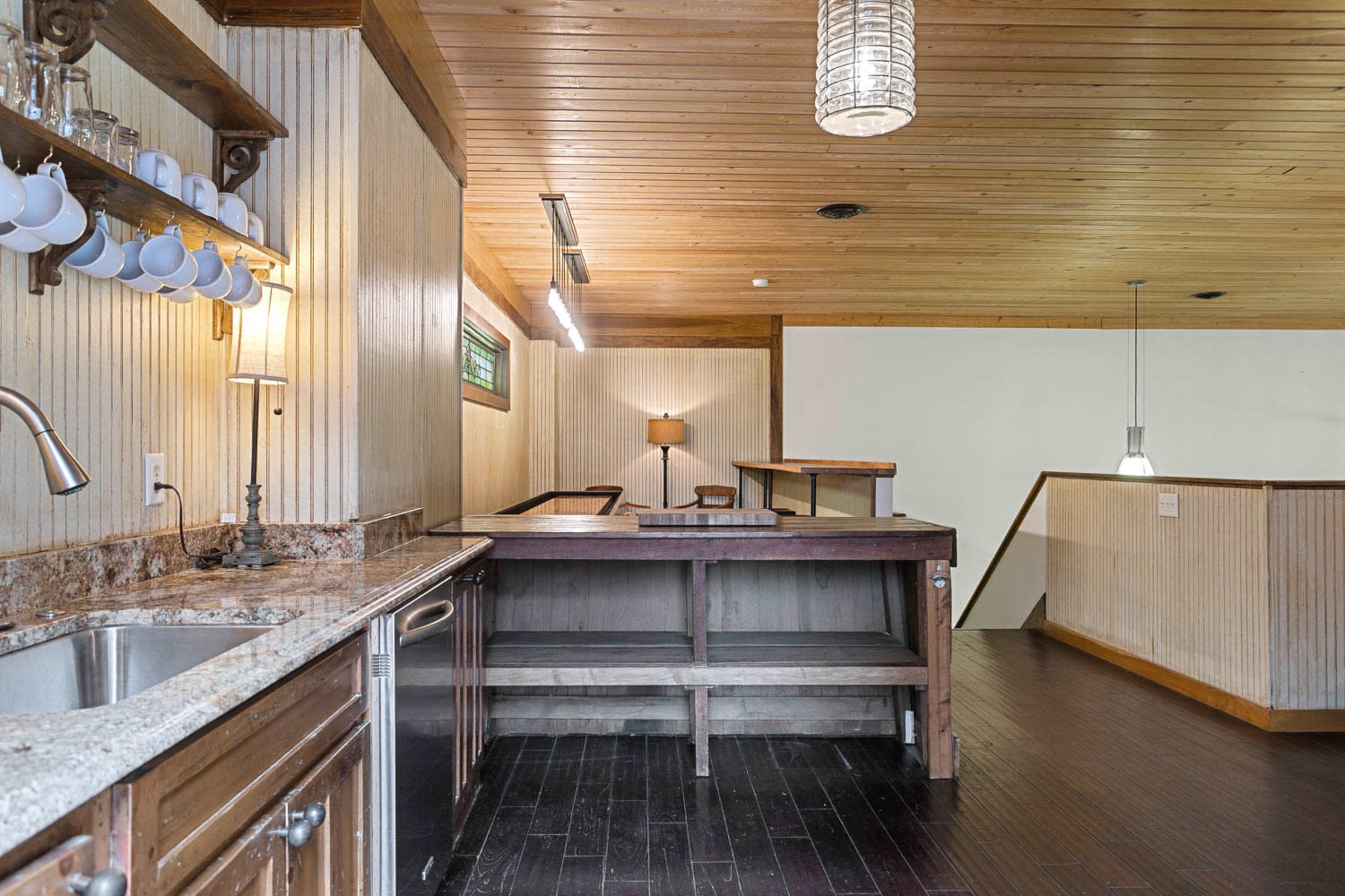 The image shows a wooden-paneled kitchen with a sink and counter area, leading to a spacious, well-lit dining or living area.