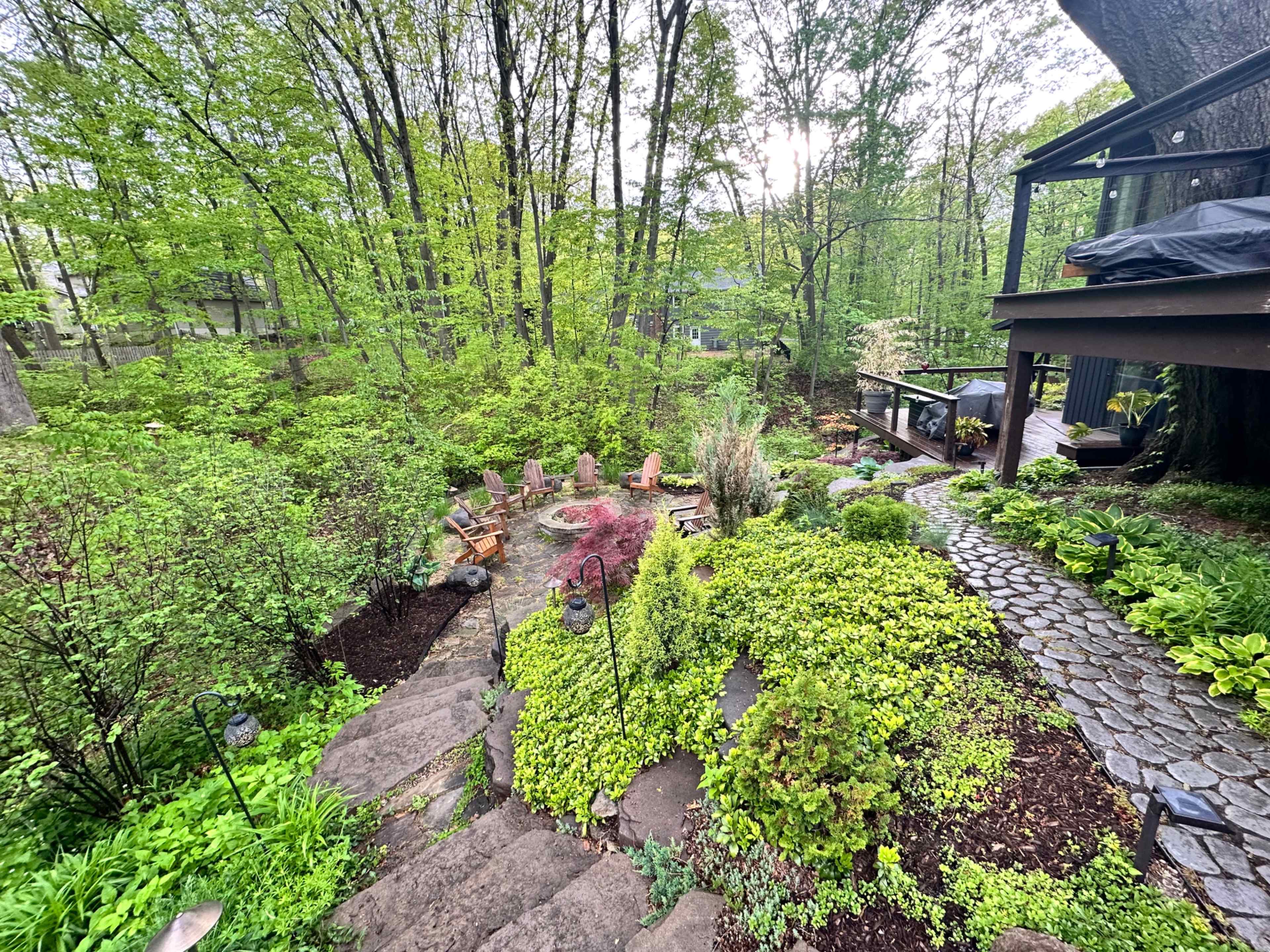 The image shows a landscaped garden with stone steps leading down to an area featuring seating, flower beds, and lush greenery, with a deck in the background.