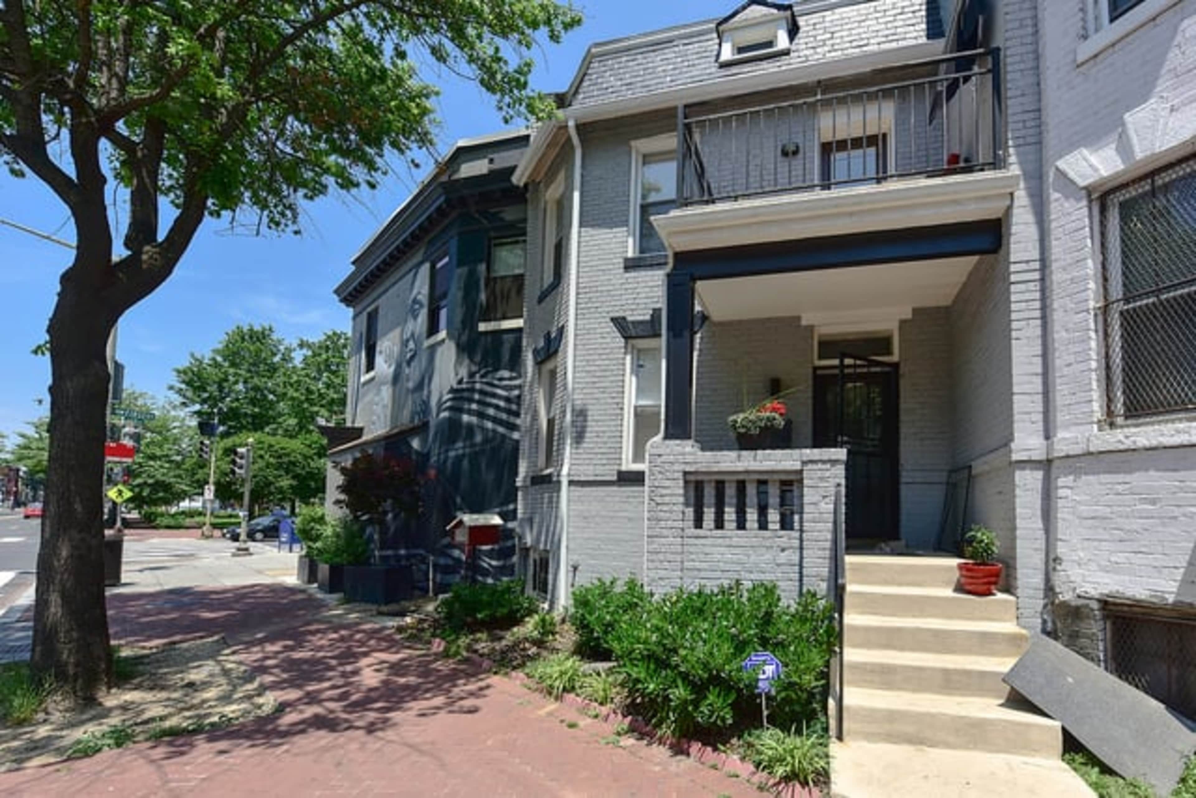 A two-story gray brick house with a front porch, flanked by greenery and a sidewalk, under a clear blue sky.