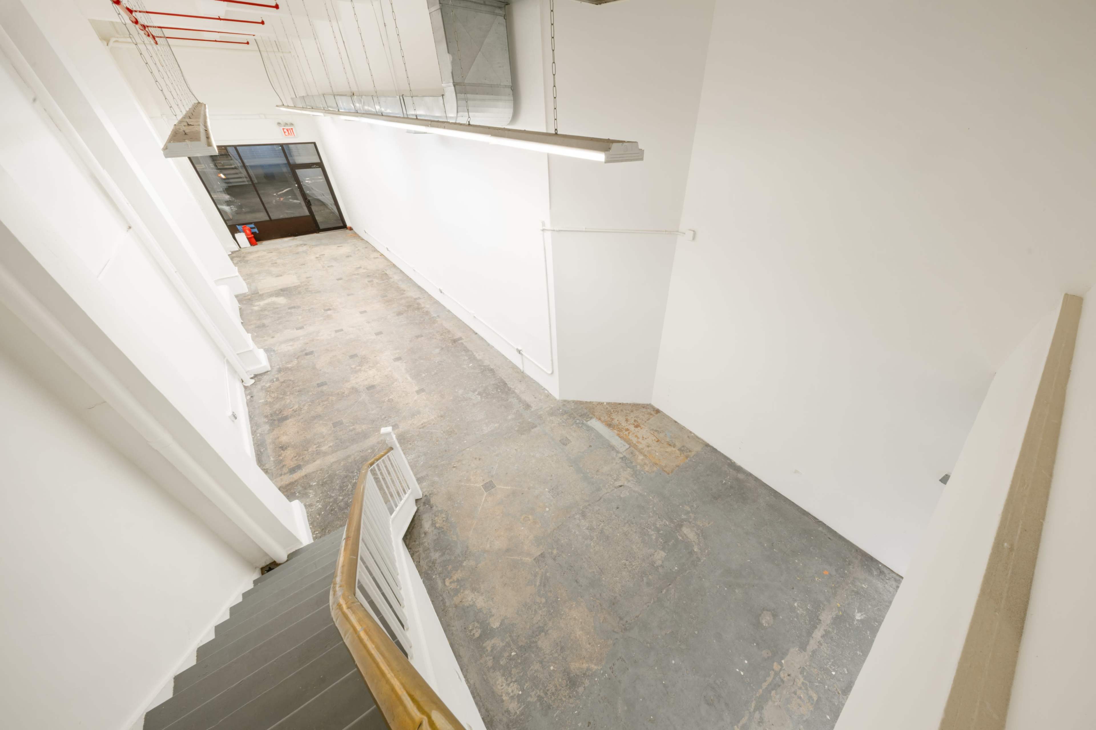The image shows a spacious, unfinished corridor with a staircase on the left and large glass doors at the far end.