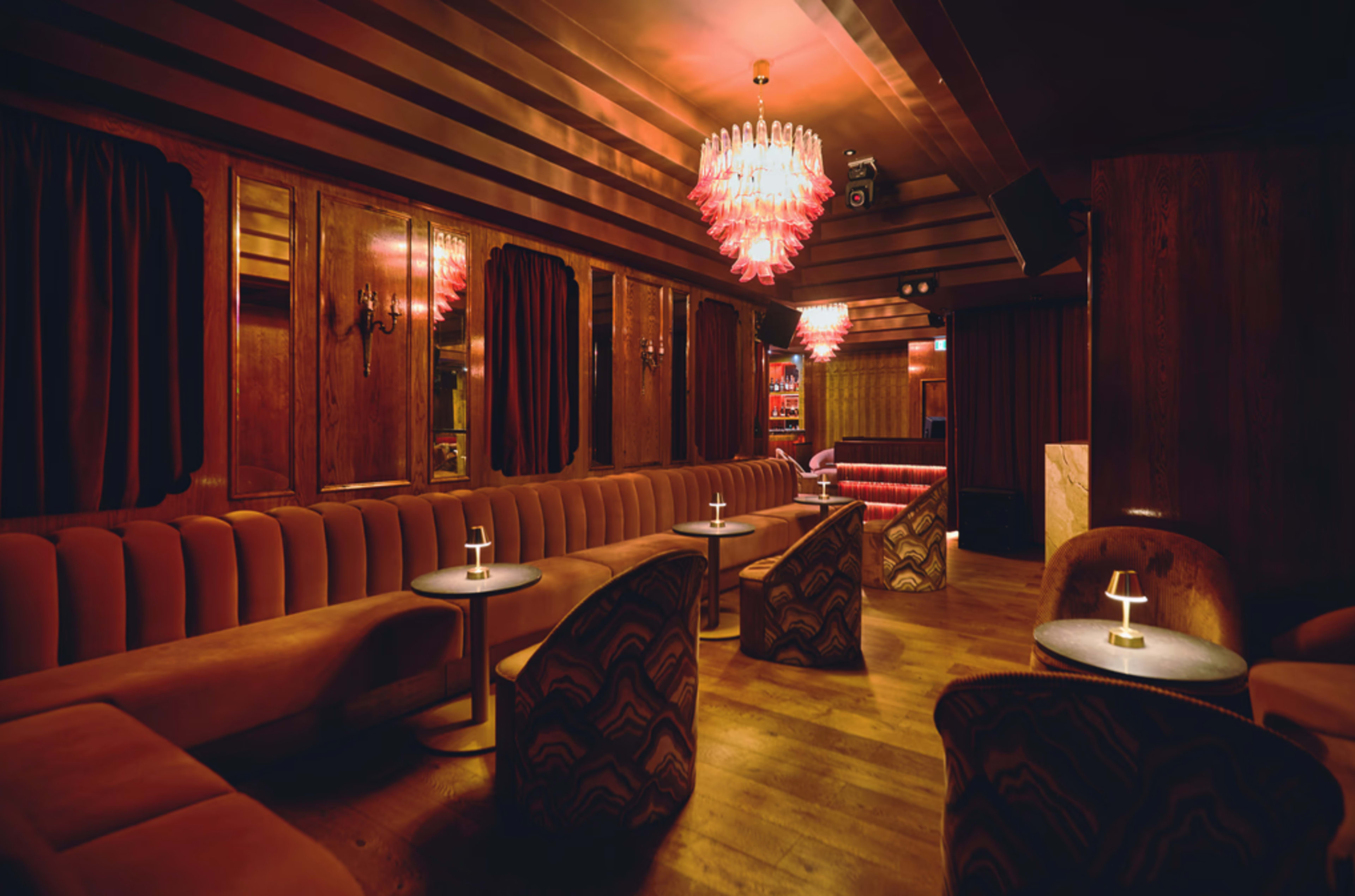 The image shows a dimly lit lounge with plush seating, wooden walls, and illuminated chandeliers.