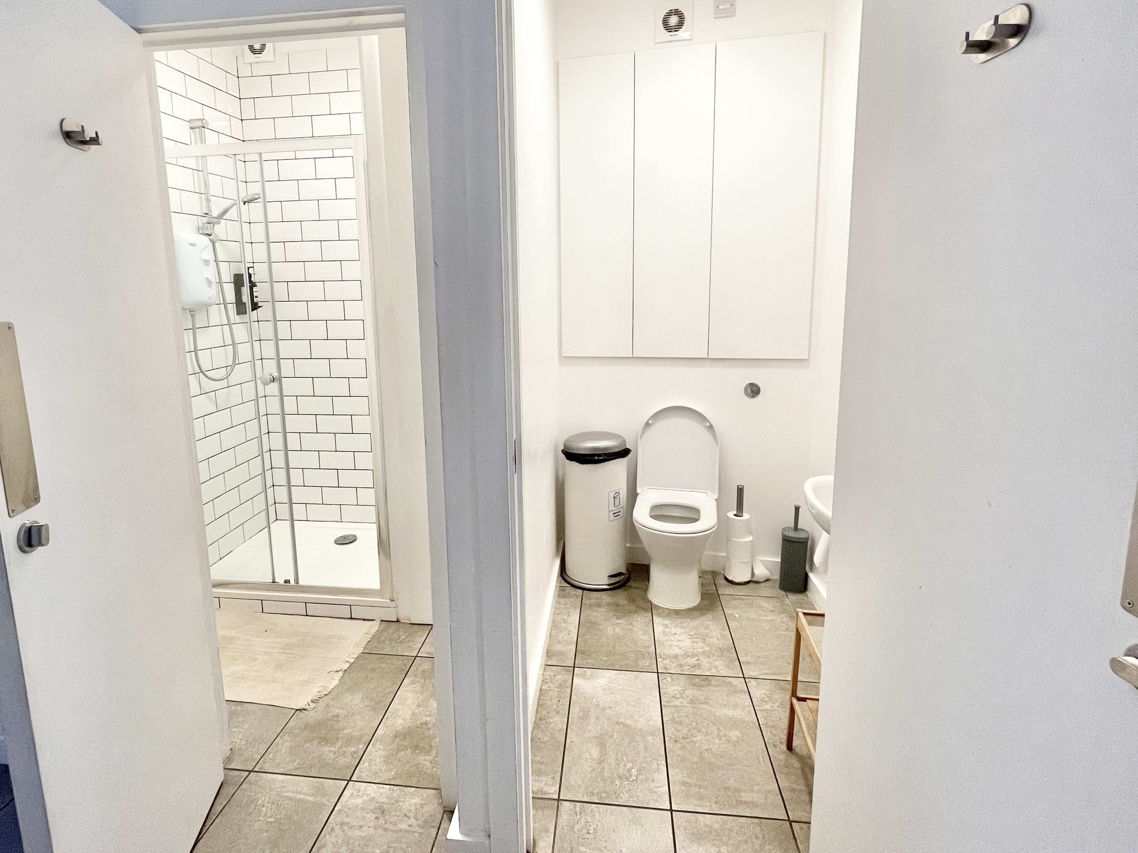 The image shows a small bathroom with a shower area on the left and a toilet and sink on the right, both sections divided by white walls.