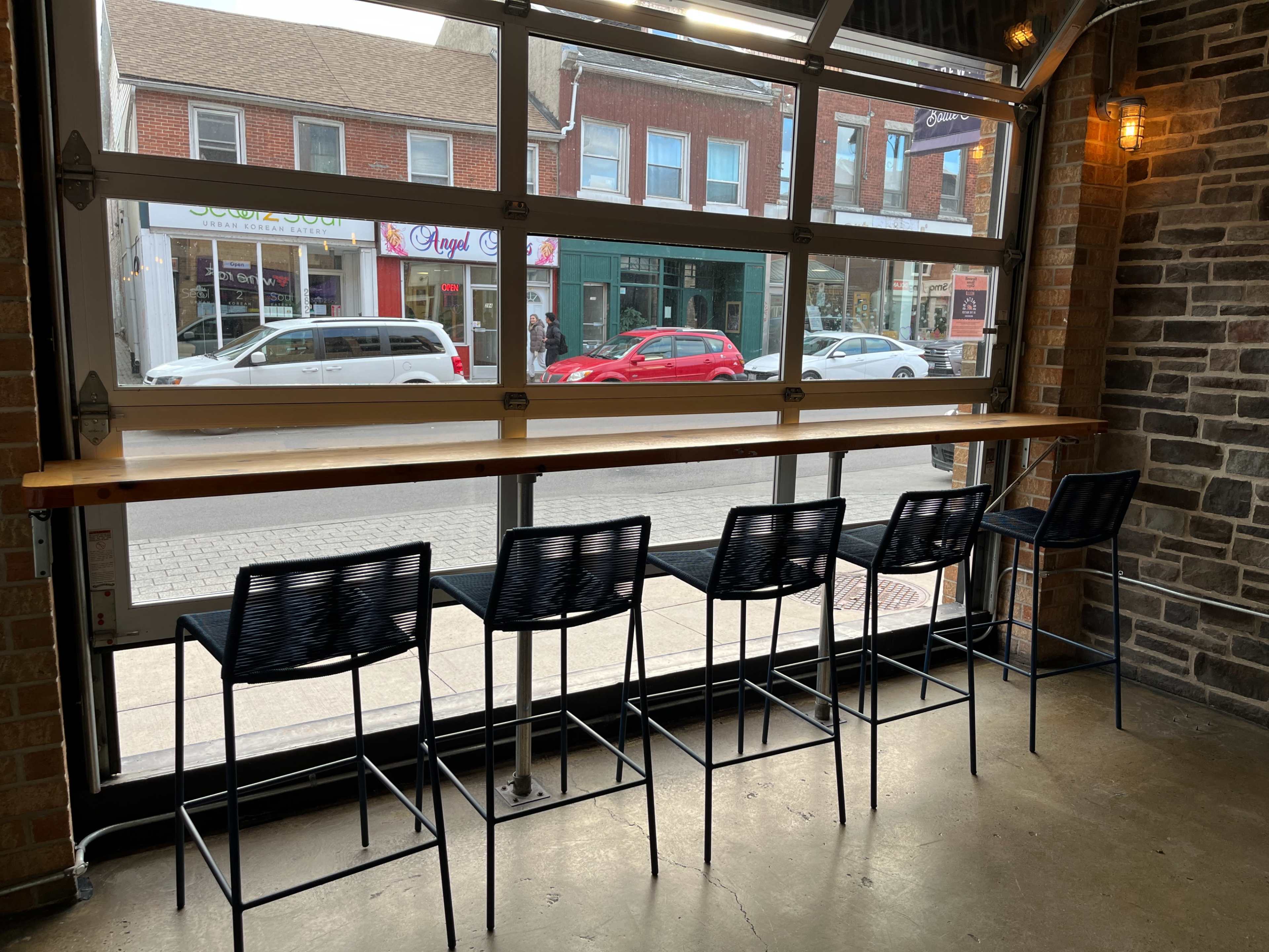 A row of five black bar stools at a wooden counter next to a large window, revealing a street view with parked cars and storefronts.