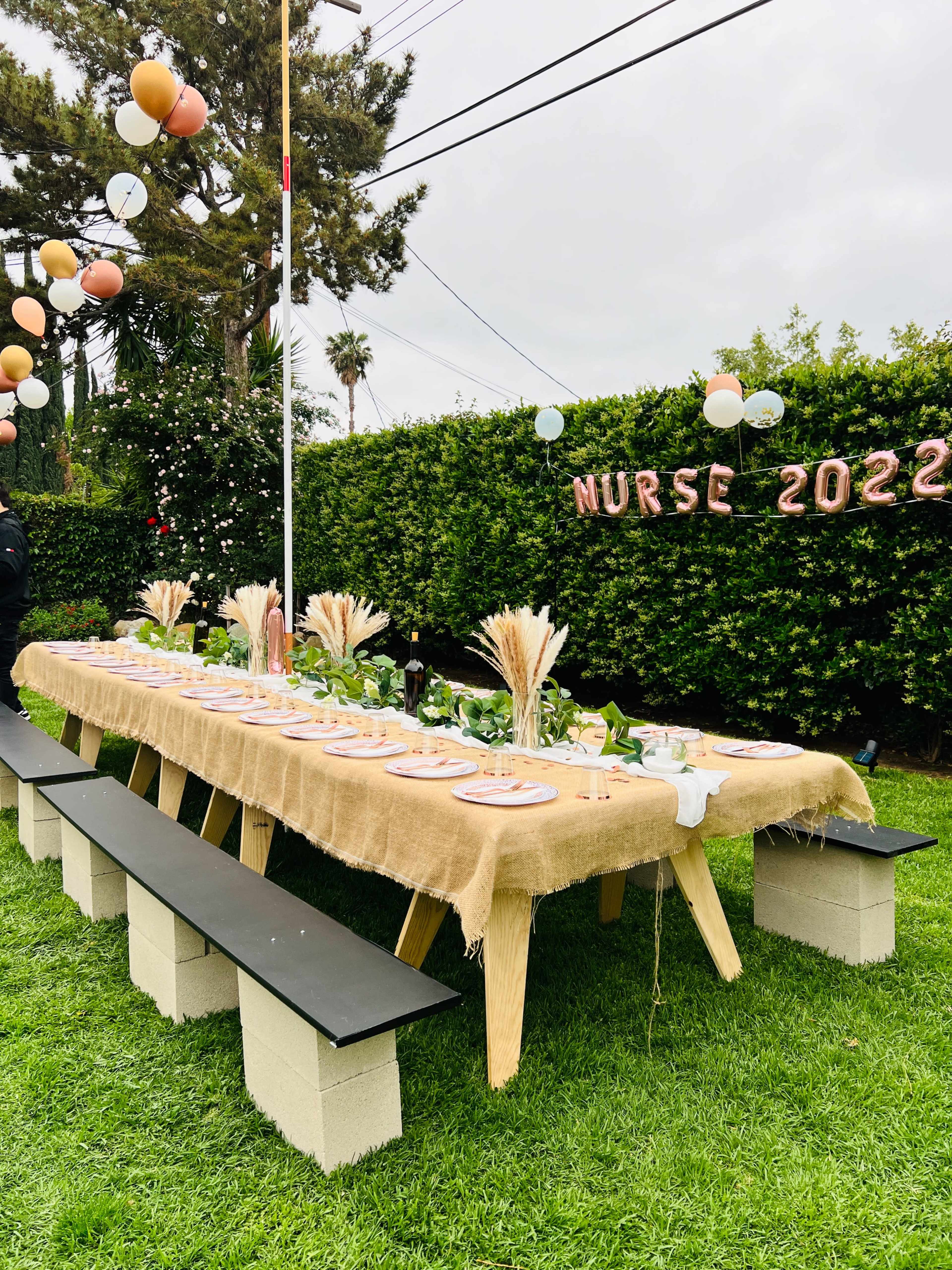 A long table set for a celebration is decorated with flower arrangements and a backdrop of greenery, with balloons and "NURSE 2022" displayed above.