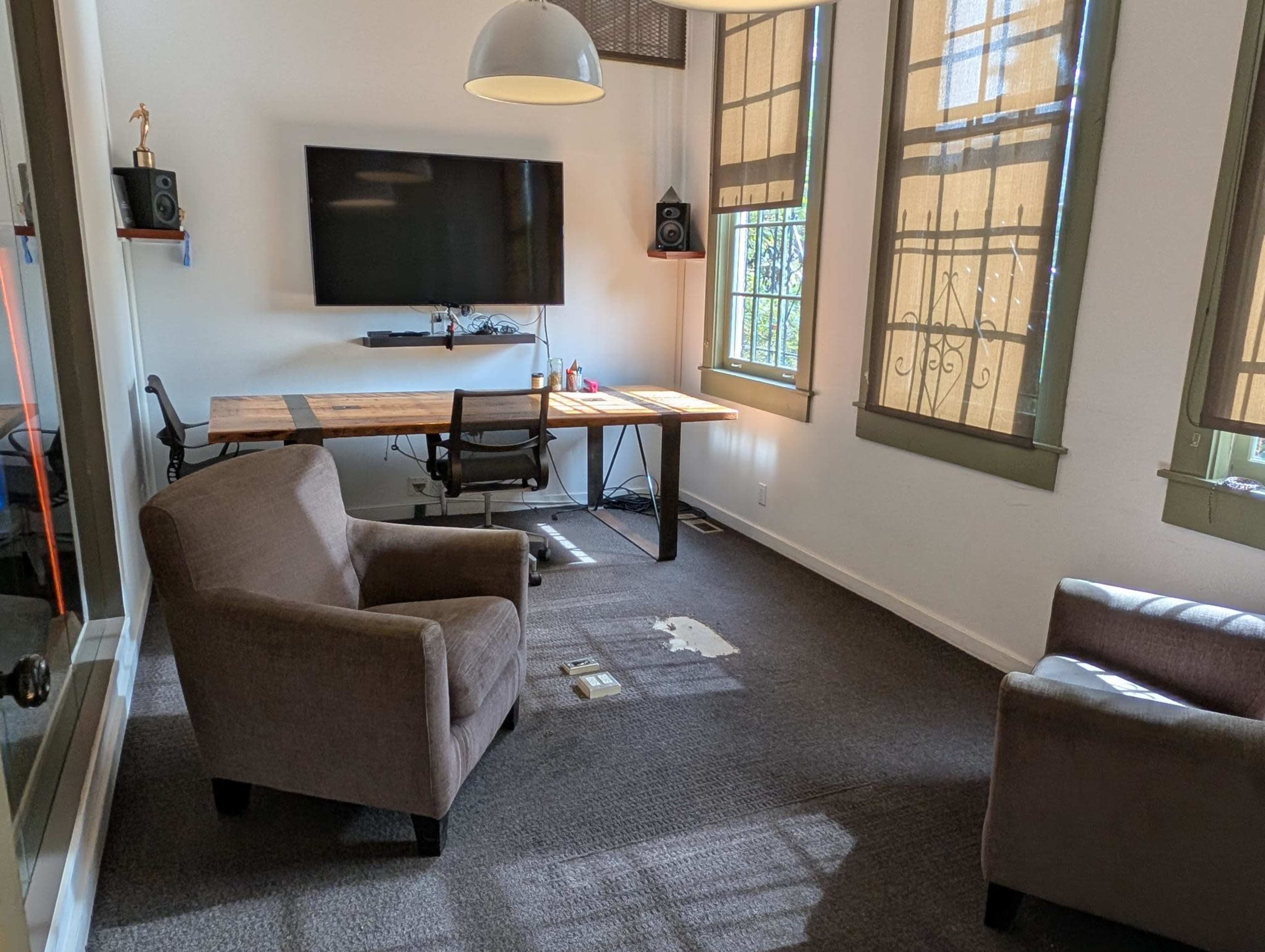 The image shows a small room with two armchairs, a wooden desk with a computer monitor, and large windows with shades.