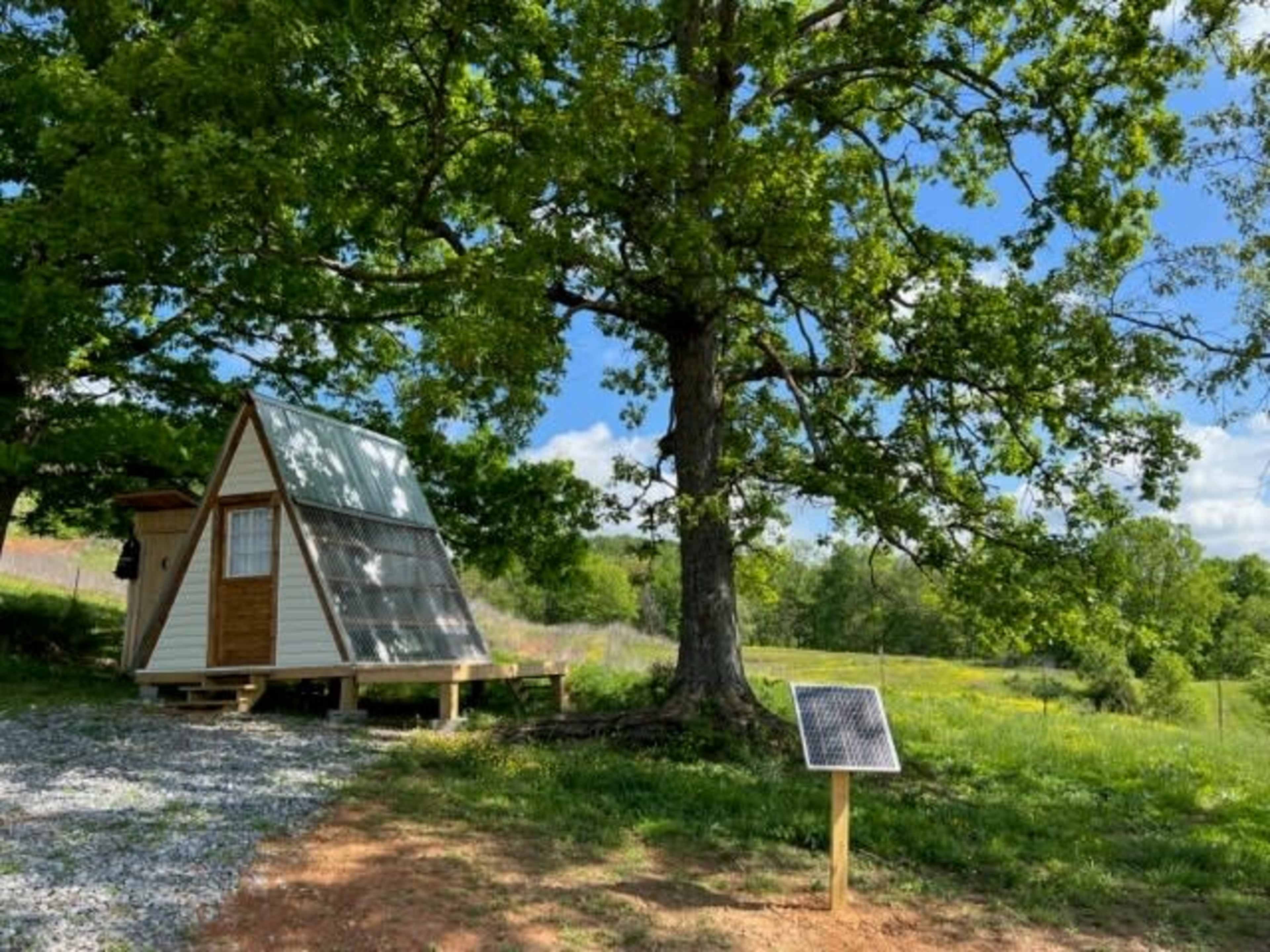 A small triangular cabin with solar panels stands beside a large tree in a grassy field.