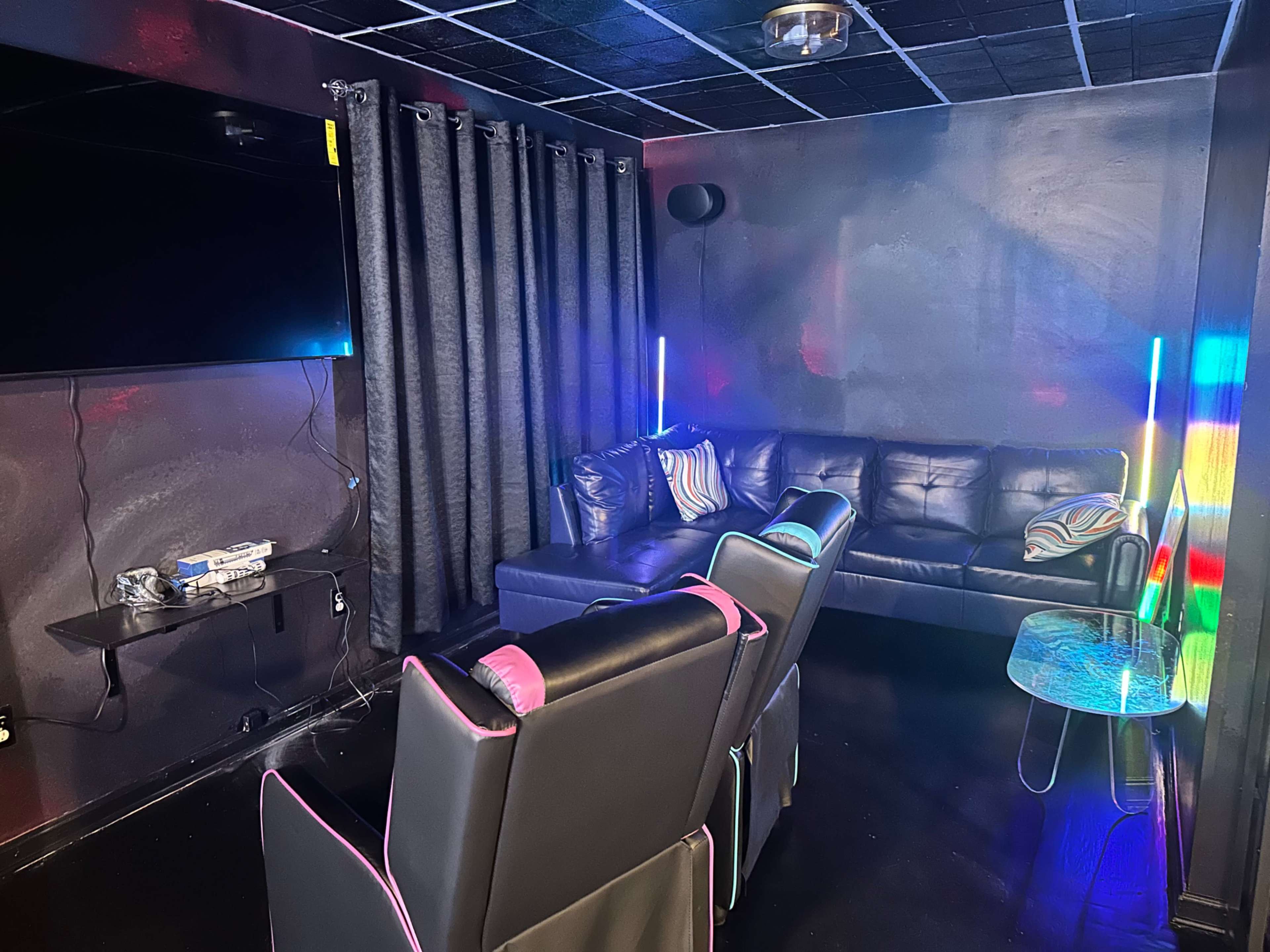 The image shows a dimly lit room featuring a black leather sofa, two gaming chairs, a small glass table, and colorful LED lights along the walls.