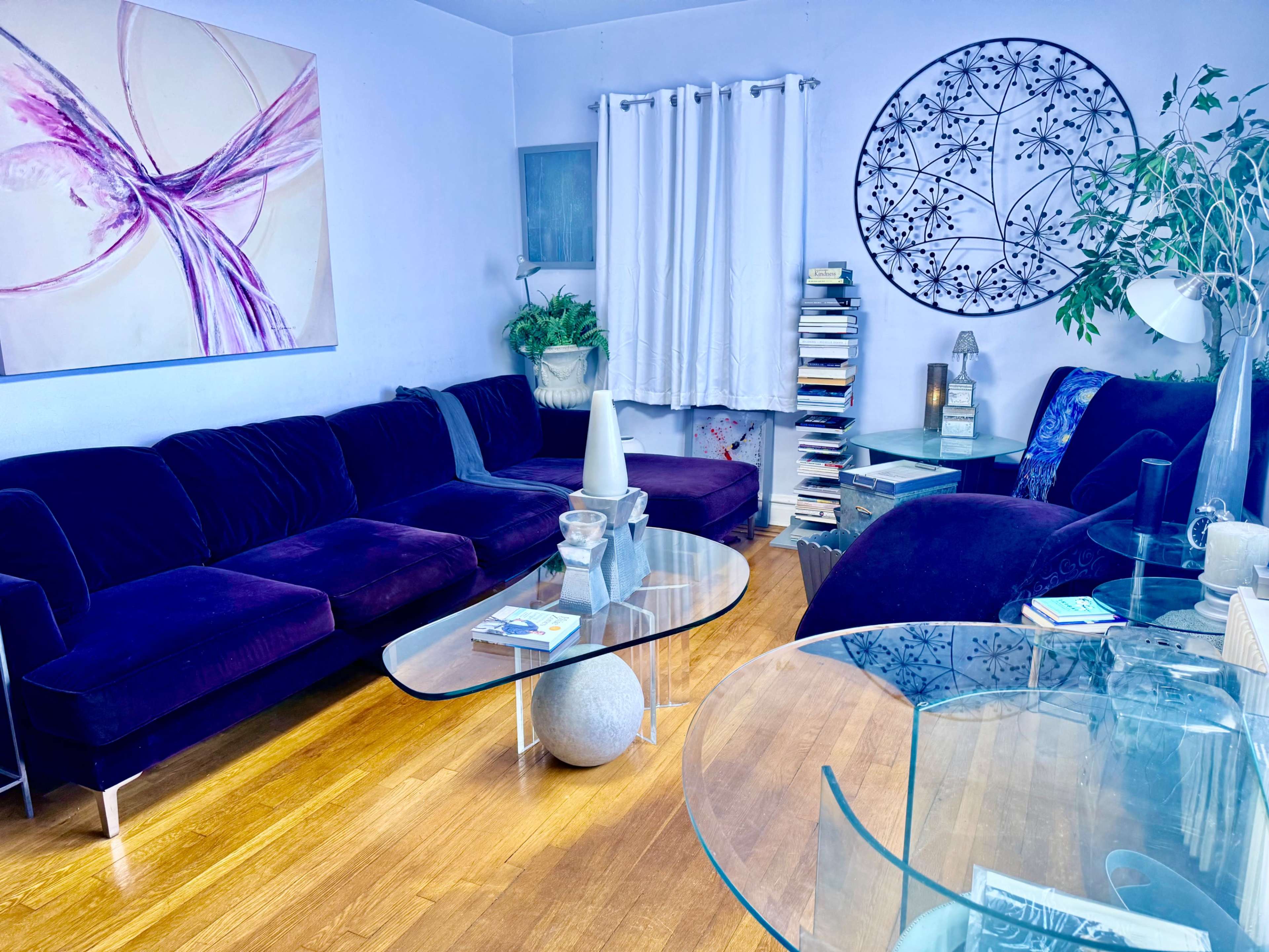 The image shows a modern living room featuring a purple velvet sectional sofa, a glass coffee table, and a circular wall art piece, with bookshelves and decorative plants in the background.