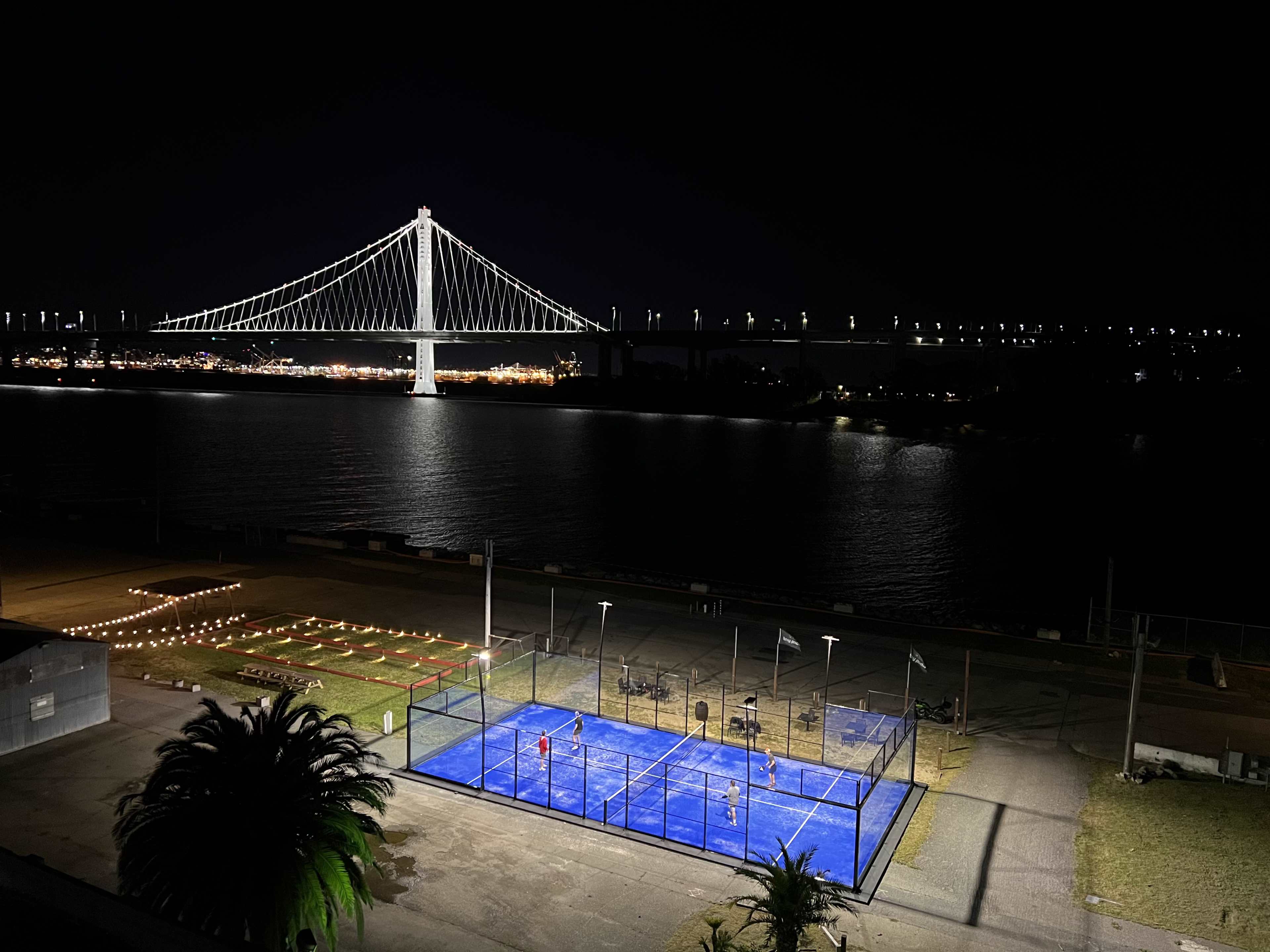 A brightly lit paddle court is positioned by the water, with a bridge illuminated in the background at night.