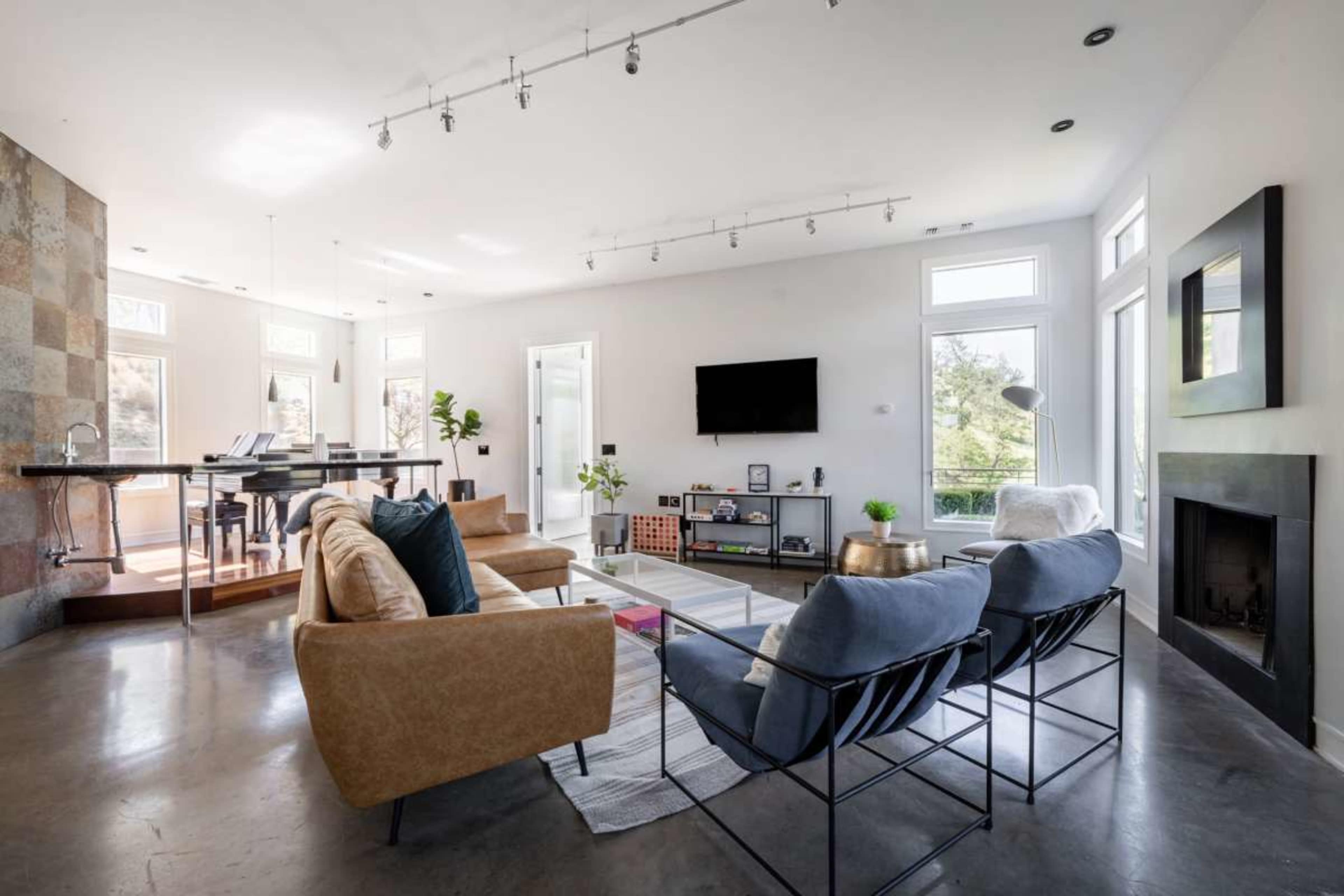 The image shows a modern living room with a mix of light-colored sofas, a television on the wall, and large windows providing natural light.